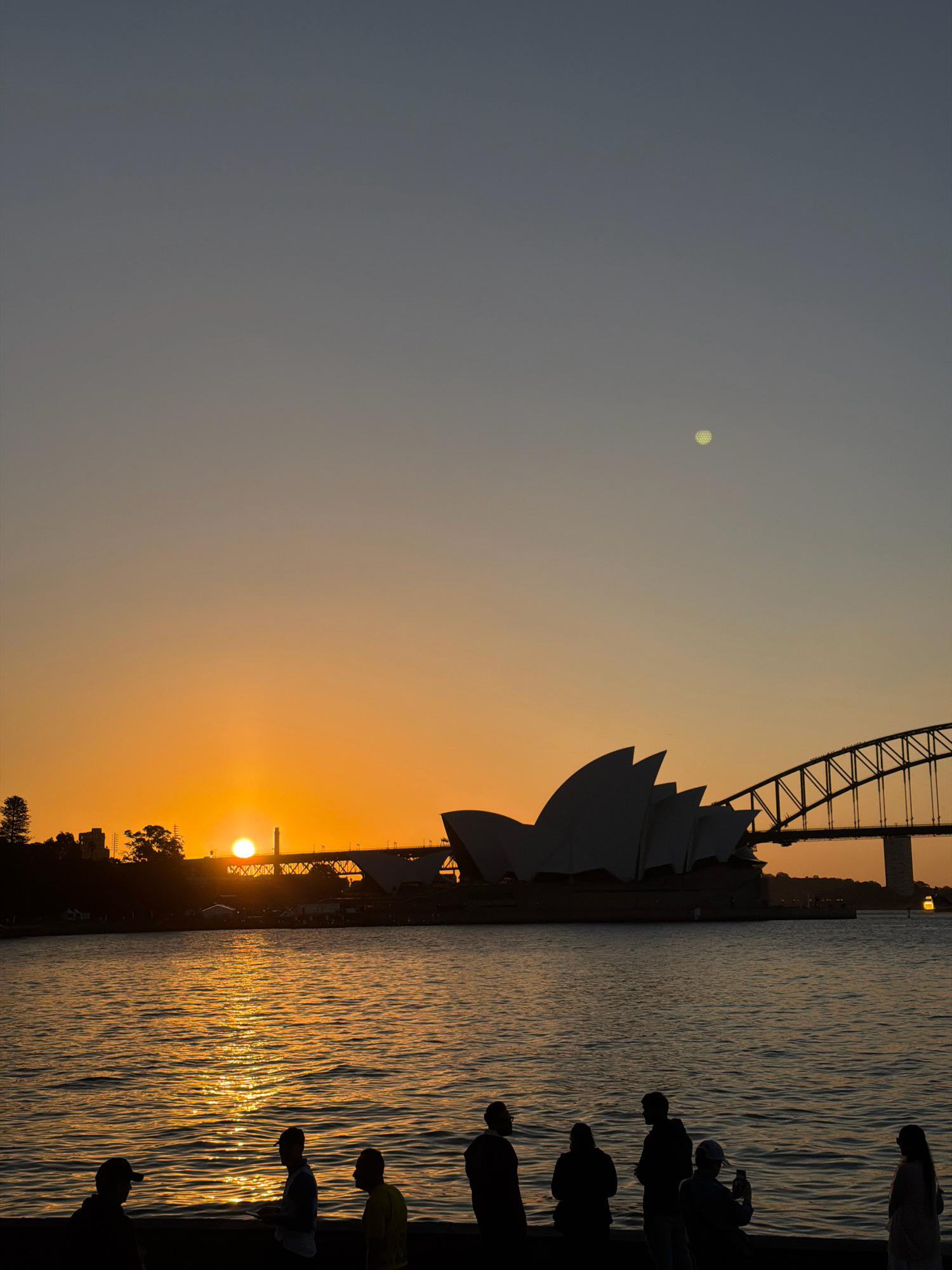 This is a vertical, silhouette photograph of the Sydney Opera House and Sydney Harbour Bridge at sunset. The sun is setting behind a bridge in the distance, casting a warm orange glow across the sky and water. The Sydney Opera House is visible in silhouette on the right, with its distinctive sail-like structures. The Sydney Harbour Bridge is also in silhouette, spanning the harbor. In the foreground, a group of people are standing on a ledge, silhouetted against the water. One person is taking a photo with their phone. The water is rippling and reflects the sunset. The moon is visible as a small, pale circle in the upper right quadrant of the sky.