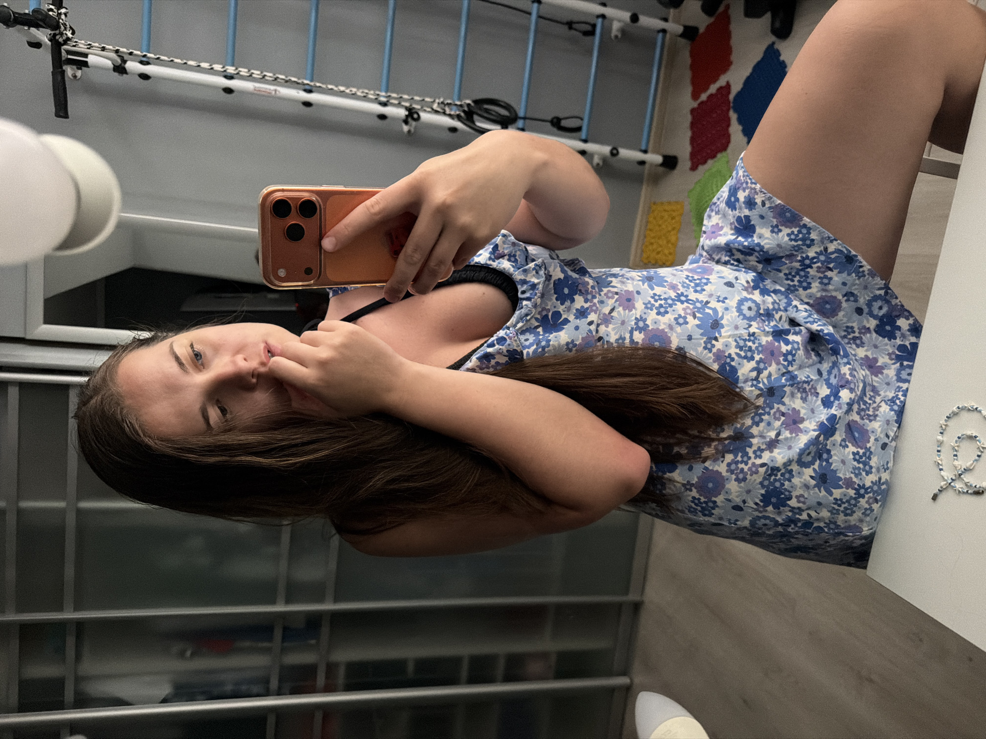 An indoor, horizontally oriented selfie captures a young woman lying down in what appears to be a residential setting in Sochi, Russia. She holds a golden/bronze smartphone with a triple camera system in her right hand, taking the picture. Her long brown hair partially obscures the left side of her face, and her left hand rests near her chin as she looks towards the camera with a casual expression.
She is wearing a blue and white floral-patterned dress or top, with a black strap visible underneath. To her right, on a white surface, lies a delicate white and blue beaded necklace or bracelet with small star motifs. The floor is covered with light wood-patterned laminate.
The background reveals several details: On the far left, a white spherical light fixture is partially visible. Further back, a white metal frame structure, possibly a drying rack or part of a home gym, extends across the upper portion of the image, featuring blue accents and woven ropes. Below this, on the floor, there's a colorful interlocking mat composed of red, blue, green, and yellow sections. Behind the woman's head, a reflective surface shows what might be vertical bars or shelving. The overall lighting suggests an indoor setting with ambient light. No discernible text is visible in the image.