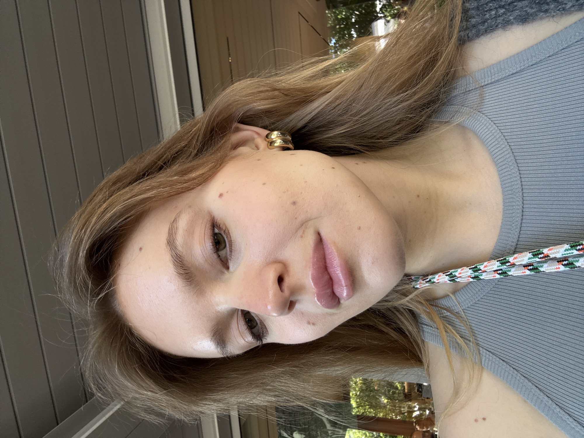 A young woman is pictured indoors. She has long, light brown hair and wears a gold hoop earring. She is facing the camera at an angle, wearing a gray ribbed tank top. A lanyard with white, green, red, and black coloring is visible around her neck. The background includes a gray slatted wall. The location in Rome, Italy, is not identifiable.