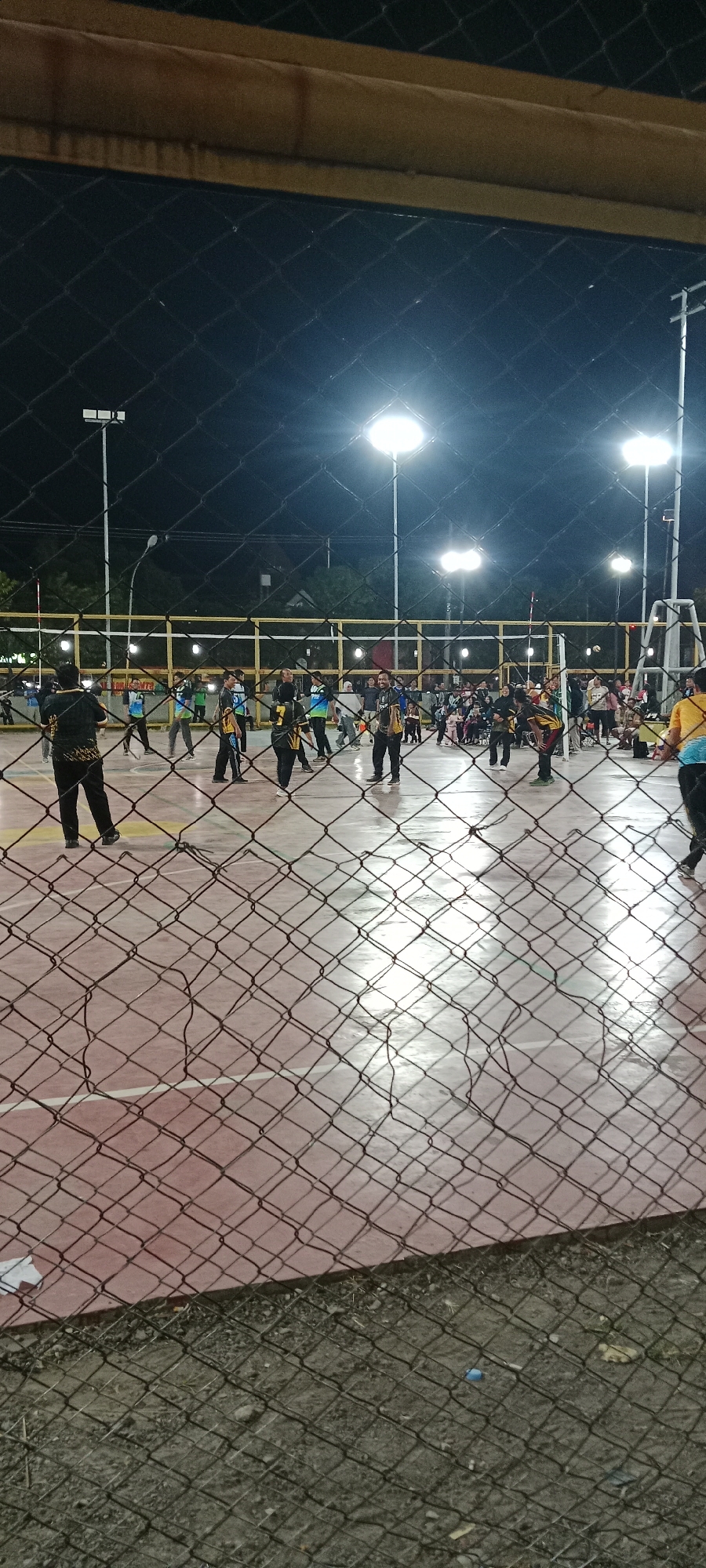 The image shows an outdoor volleyball court at night, illuminated by floodlights. A group of people are present on the court, some of whom appear to be playing volleyball. The court surface is a reddish-orange color. A chain-link fence is in the foreground, partially obscuring the view of the court. In the background, there are trees and buildings, suggesting an urban or suburban setting. The location is identified as Tuban, Indonesia.
