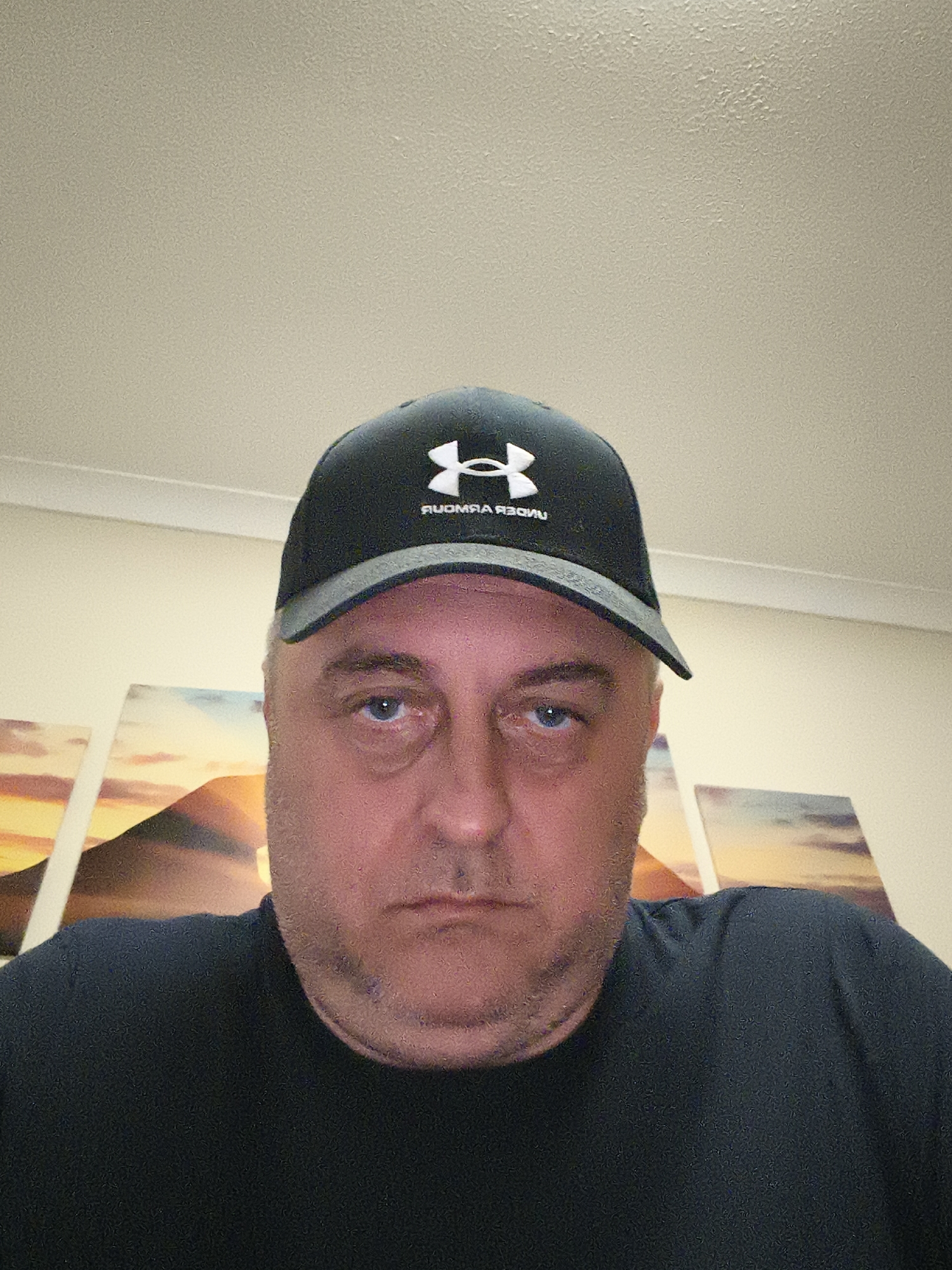 The image features a middle-aged man in a close-up, head-and-shoulders shot, looking directly at the camera. He has blue eyes, a light complexion, and visible stubble. He wears a dark-colored baseball cap with a grey brim, displaying the reversed text "ЯUOMЯA ЯƎGNU" (Under Armour) and its logo. He is also wearing a plain, dark-colored crew-neck t-shirt.
The man is indoors, with a neutral to slightly somber expression. The background includes a light-colored, textured ceiling and a wall behind him adorned with at least three rectangular artworks or prints. These artworks depict a vibrant landscape with warm tones of orange, yellow, and blue, possibly illustrating a sunset or sunrise over mountains. The uniform lighting suggests an indoor artificial light source. The reversed text on the cap is a notable detail, indicating the image is likely a selfie. The external location context provided is Huntingdonshire, United Kingdom.