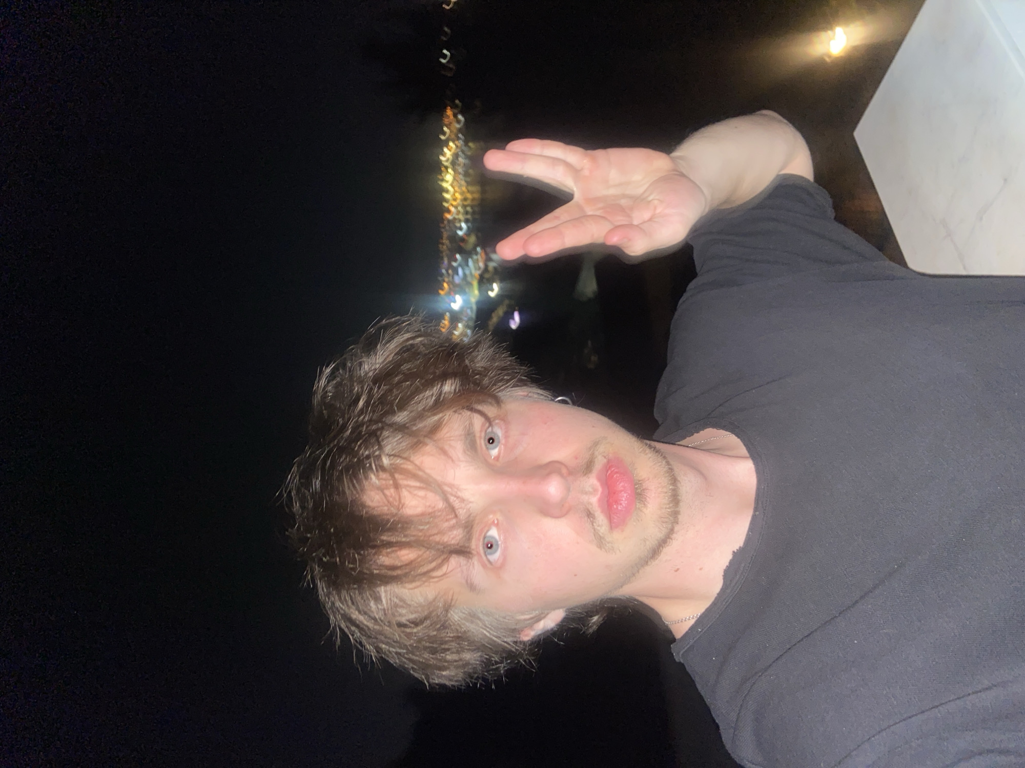 A young man is photographed at night in what appears to be Šušanj, Montenegro, indicated by blurred nighttime city lights in the background. The man, positioned in the foreground, has disheveled brown hair, pale skin, and light-colored eyes. He has a light beard and mustache. He is wearing a dark-colored, possibly black, t-shirt. The man is making a hand gesture with his right hand; two fingers are extended upward, forming a "peace sign". A white surface or object is visible in the upper right corner. The overall lighting is dark, likely illuminated by distant city lights.