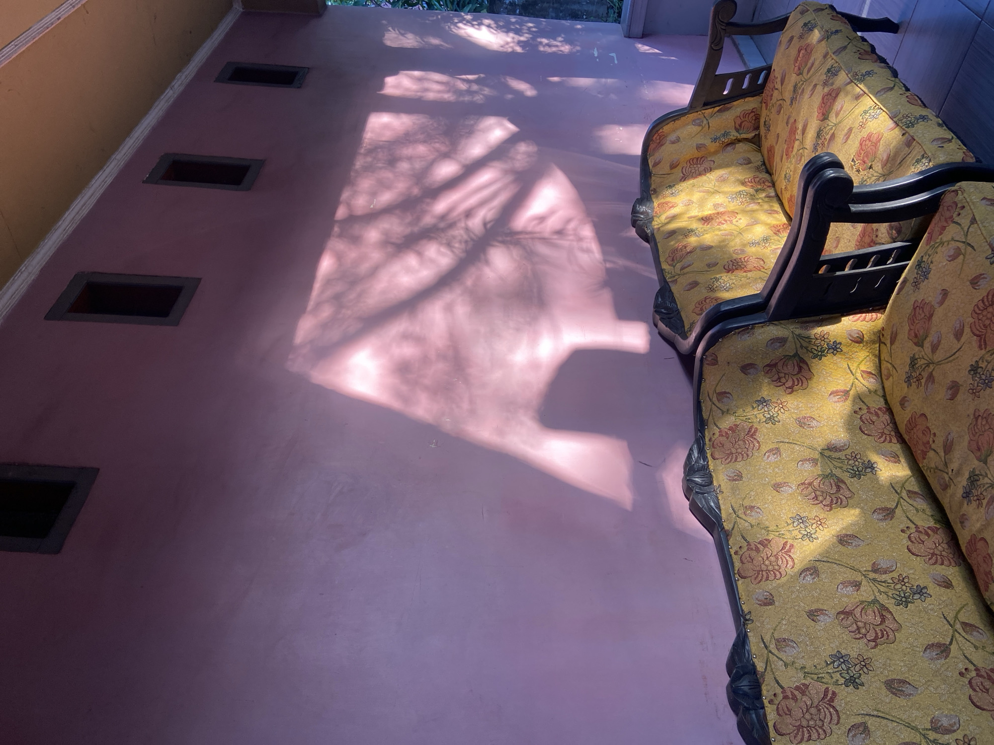 The image depicts a sunlit porch area with two vintage upholstered sofas positioned against a textured pink wall. The sofas have dark wood frames and are covered in a yellow fabric adorned with a floral pattern of red and pink roses with green leaves and smaller blue and white flowers. Sunlight casts a dappled shadow of tree branches across the pink wall, creating a dynamic interplay of light and shade. The wall has several small, rectangular vents or windows near the top, and a white decorative trim runs along the ceiling line, which is a warm, muted orange. The floor appears to be tiled. The setting suggests an outdoor residential space in a tropical or subtropical climate. The image does not contain any people or explicit indications of a specific location within Palopo, Indonesia, beyond the implied residential setting.
