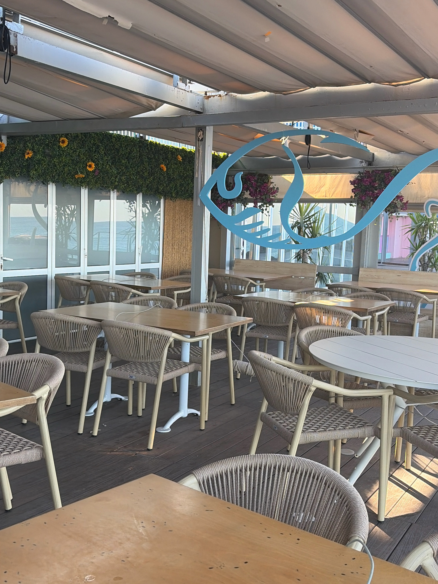 The image depicts an outdoor seating area of a restaurant or cafe in Salou, Spain. The establishment appears to be situated near the coast, as evidenced by the view of the sea visible through large glass doors in the background. The space is furnished with numerous tables and chairs, primarily constructed from woven rattan-like material and light-colored metal frames, arranged on a wooden deck. Some tables are round and white, while others are rectangular with wooden tops. Hanging above the seating area is a retractable awning, offering shade. The decorative elements include a large blue fish outline graphic on a glass partition and artificial greenery with yellow flowers along a railing. The overall ambiance suggests a relaxed, possibly beachside dining experience. There are no people visible in the scene.