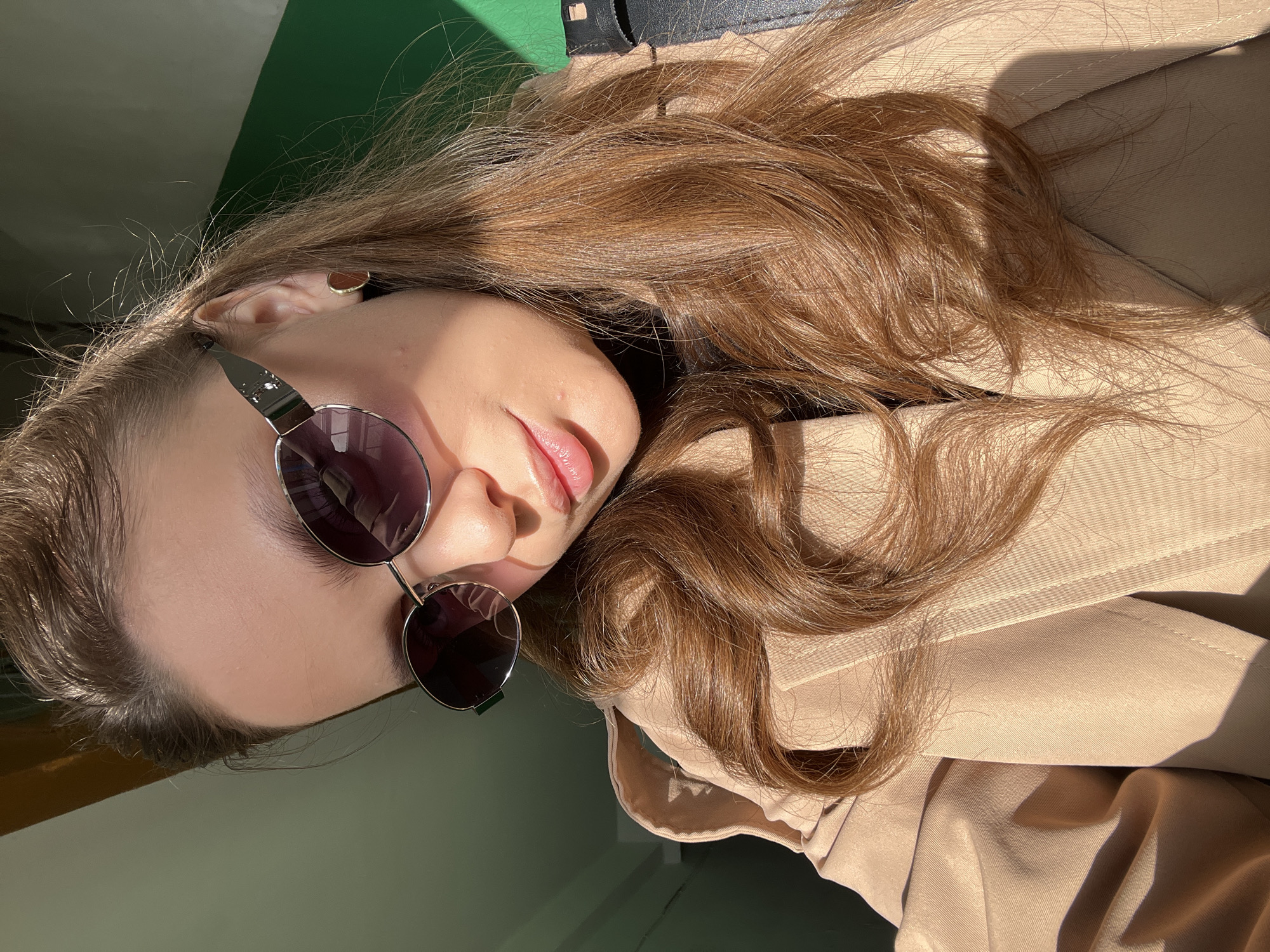 A fair-skinned person with long, wavy brown hair is lying on a beige-colored garment, possibly a jacket or coat. The person is wearing dark, round sunglasses with a silver frame and a small, circular earring. The background is divided into two color sections: a muted green at the bottom and a light green at the top. A strip of what appears to be black fabric is visible near the top. The lighting indicates an outdoor or sunlit location, casting shadows on the garment and the person's face. The photograph appears to be taken at an angle. It's not possible to determine the location as Chaikovsky, Russia from the image.