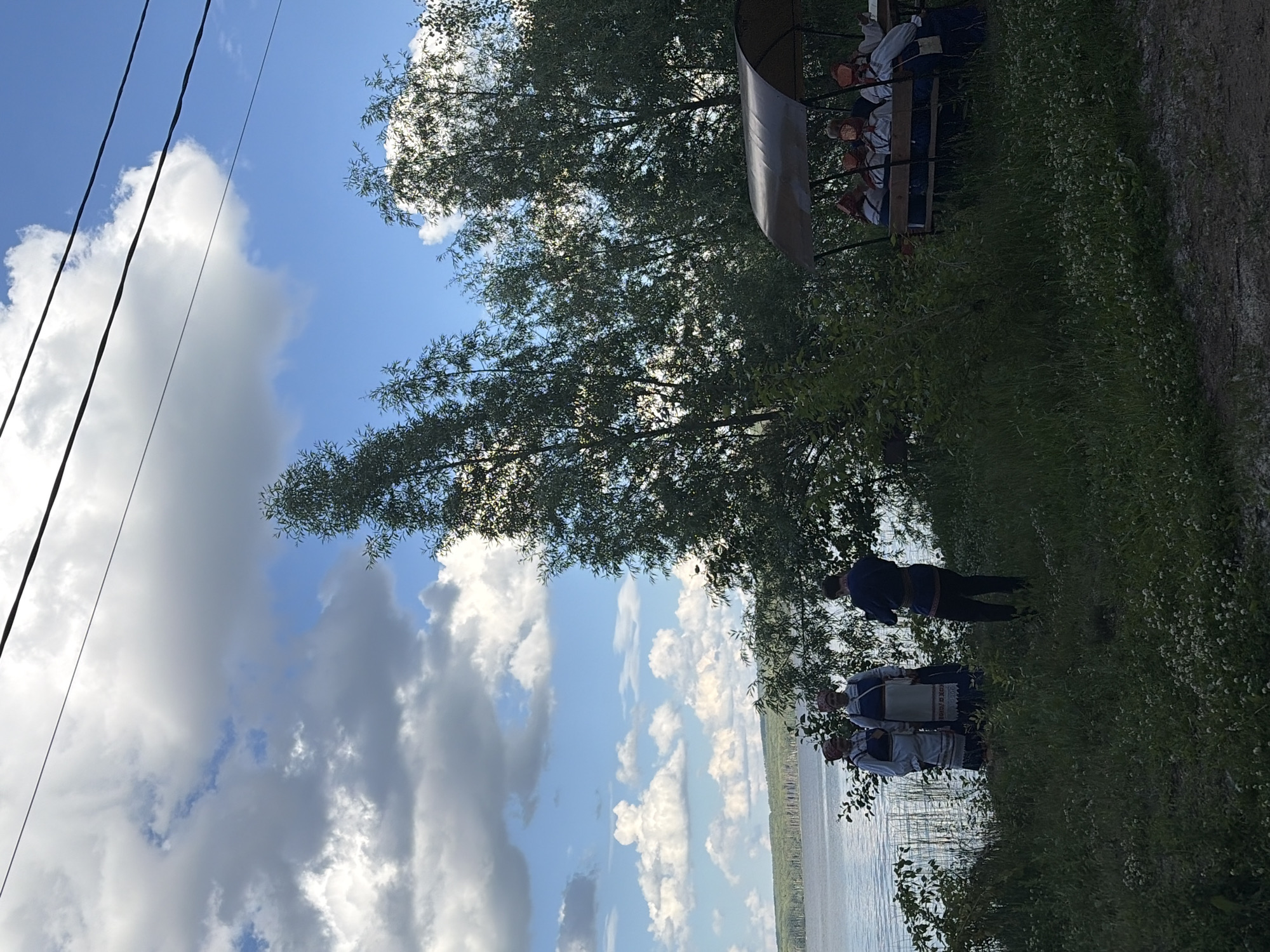 The image depicts a lakeside scene in Chaikovsky, Russia. In the foreground, on a grassy embankment, three individuals are present. Two women, dressed in traditional folk costumes featuring embroidered blouses and headscarves, stand together. A third individual, a man in a dark blue patterned shirt and dark trousers, stands behind them, facing away from the viewer. To the right, a covered seating area with a dark brown roof is occupied by several people, also appearing to be dressed in folk attire, facing inwards. They are seated on a wooden bench. The background features a calm body of water, likely a reservoir given the location, with a treed shoreline and a distant, hazy horizon. The sky is a mix of blue sky and large, fluffy white and grey clouds. Several dark wires are visible stretching across the upper part of the image. The overall atmosphere suggests an outdoor gathering or event, possibly cultural or historical, taking place on a bright, cloudy day near a body of water.