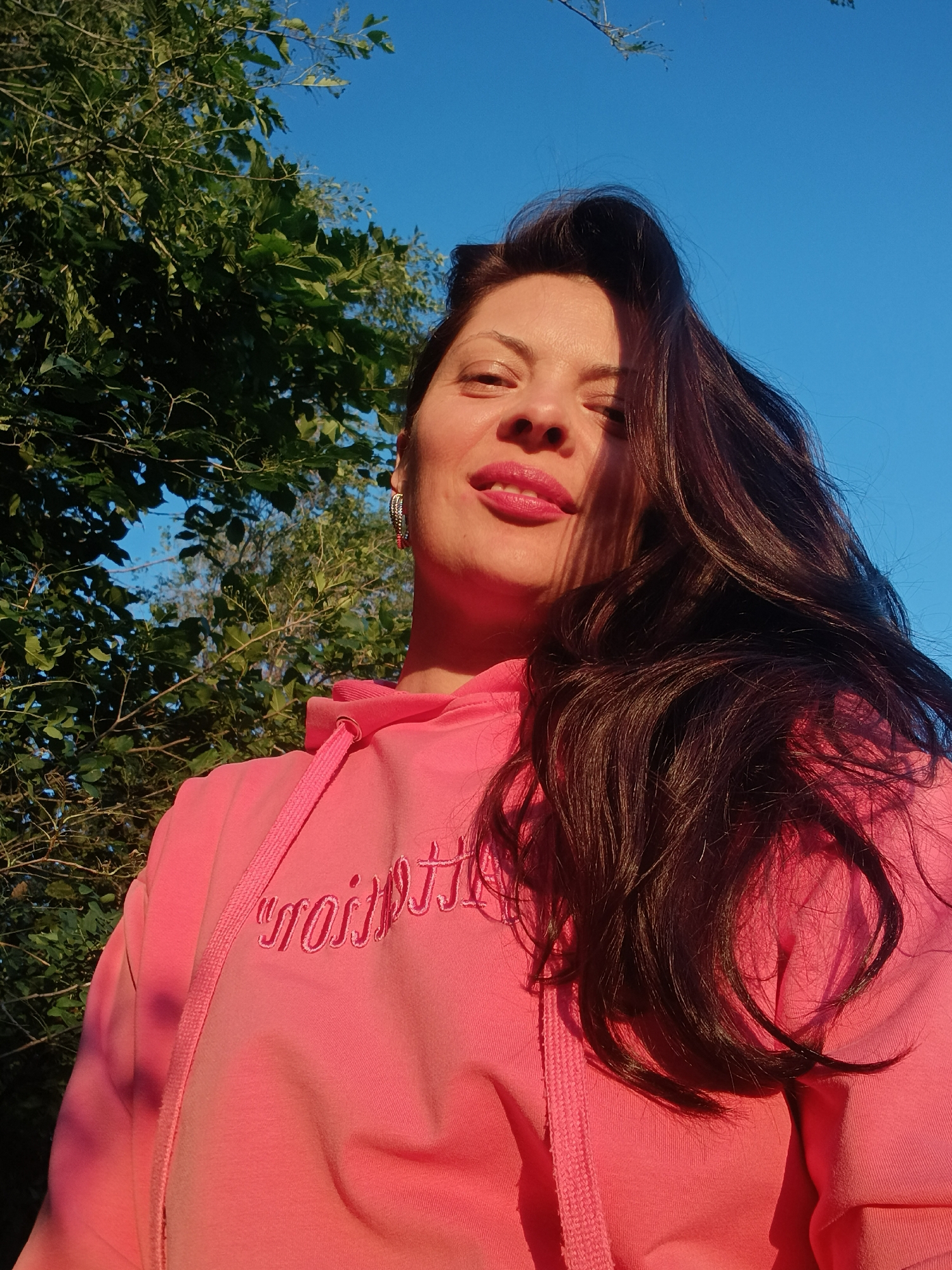A woman, with dark hair and bright pink lipstick, poses for a selfie outdoors under a clear blue sky. She is wearing a pink hooded sweatshirt with "Lollitop" embroidered in a cursive font across the chest. Green foliage from trees is visible in the background, along with a hint of what appears to be a building. The lighting suggests it is late afternoon or early evening.