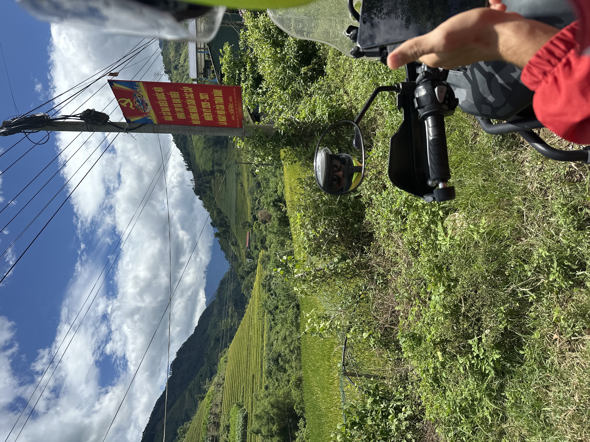 The image captures a first-person perspective from a motorcycle, depicting a rural or mountainous landscape in Nam To Nua, Vietnam. In the immediate foreground on the right, parts of a motorcycle are visible, including a black handlebar with a handguard, a side mirror reflecting the rider's face (wearing dark sunglasses and a helmet with a yellow-green trim), and a section of a camouflage-patterned motorcycle body. A hand, covered by a red sleeve, is partially visible on the right, positioned near the handlebars.
The midground is dominated by lush green vegetation, including various grasses, shrubs, and small trees. A utility pole stands prominently on the right, supporting multiple black overhead wires that traverse the sky. Attached to this pole is a vertical red banner. The banner features a yellow hammer and sickle emblem with a star at the top, alongside Vietnamese text. The text reads: "BẦU CỬ ĐẠI BIỂU QUỐC HỘI KHÓA XVI VÀ ĐẠI BIỂU HỘI ĐỒNG NHÂN DÂN CÁC CẤP NHIỆM KỲ 2026 - 2031 LÀ NGÀY HỘI CỦA TOÀN DÂN!" (Election of National Assembly Delegates XVI Term and Delegates People's Councils at all levels Term 2026 - 2031 Is the festival of all the people!).
The background features steep, verdant mountains covered in dense foliage. On the slopes of these mountains, distinctly terraced agricultural fields, appearing light green, are visible. Further in the distance, additional mountains are discernible through a bluish atmospheric haze. A structure with a red roof is partially visible among the trees on a distant slope. The sky above is a bright blue, interspersed with scattered white cumulus clouds.