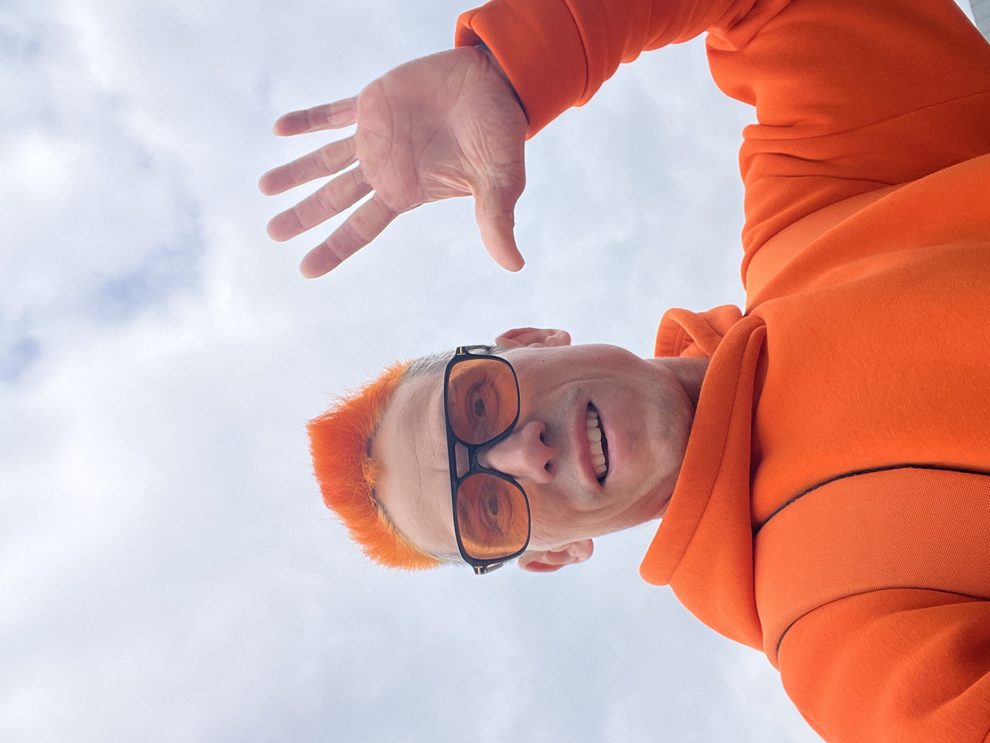 A man wearing an orange hooded sweatshirt and orange-tinted sunglasses is looking down at the camera. He has orange hair and his right hand is raised in front of his face. The background is a cloudy sky. The location is unknown.