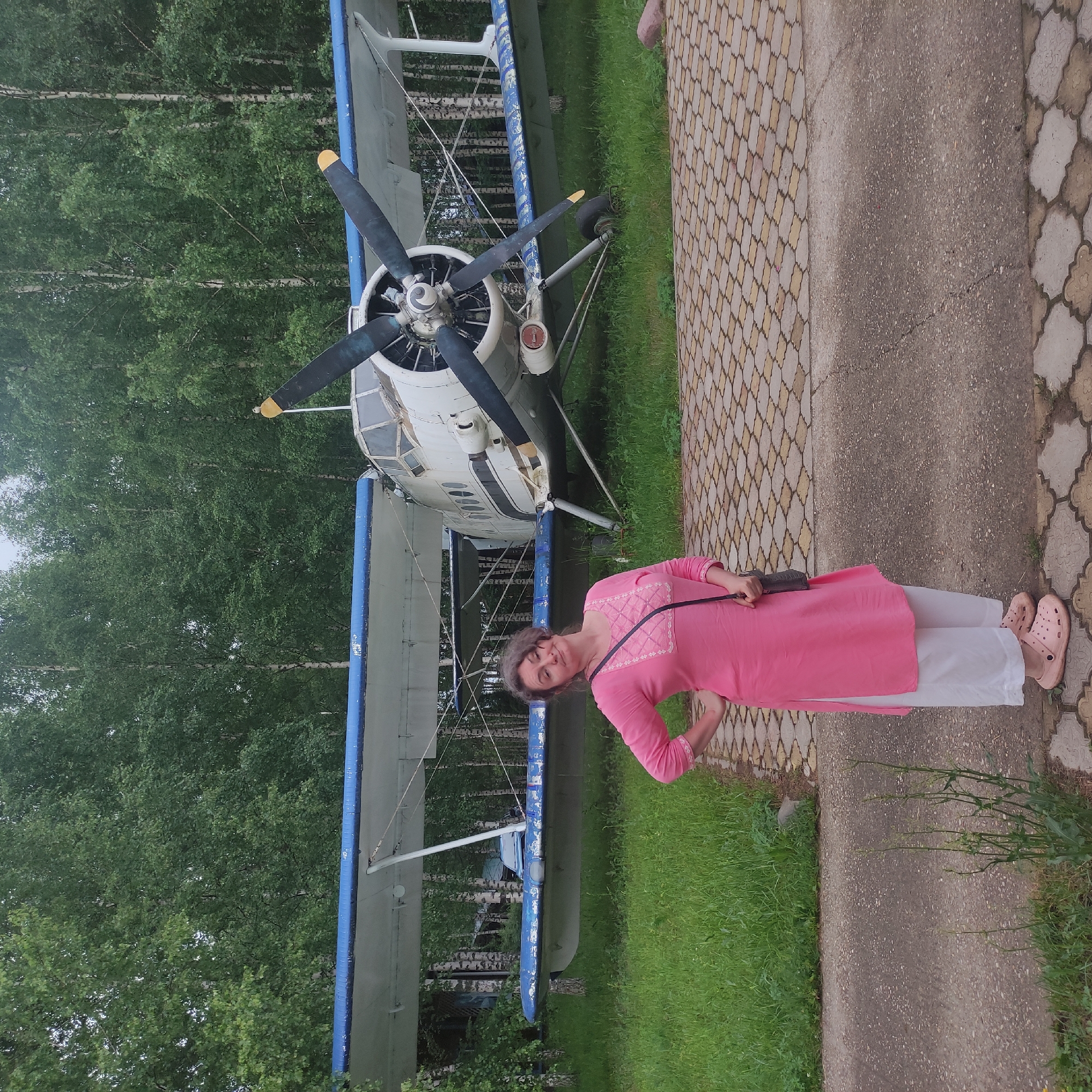 A woman with curly brown hair stands in front of a vintage biplane parked on a grassy area. She is wearing a bright pink tunic with white embroidery on the neckline and sleeves, white pants, and pink clogs. A dark crossbody bag is slung over her right shoulder. Her left hand is on her hip, and her head is tilted slightly to her left, with a playful expression. The biplane is white with blue trim on its wings and tail. It has two propellers and appears to be weathered. Behind the plane, a dense forest of birch trees with white trunks and green foliage is visible under an overcast sky. The ground in front of the plane is paved with interlocking stone tiles. The scene is likely outdoors, possibly in a museum or historical site dedicated to aviation, in or near Zhirnosovo, Russia, given the context.