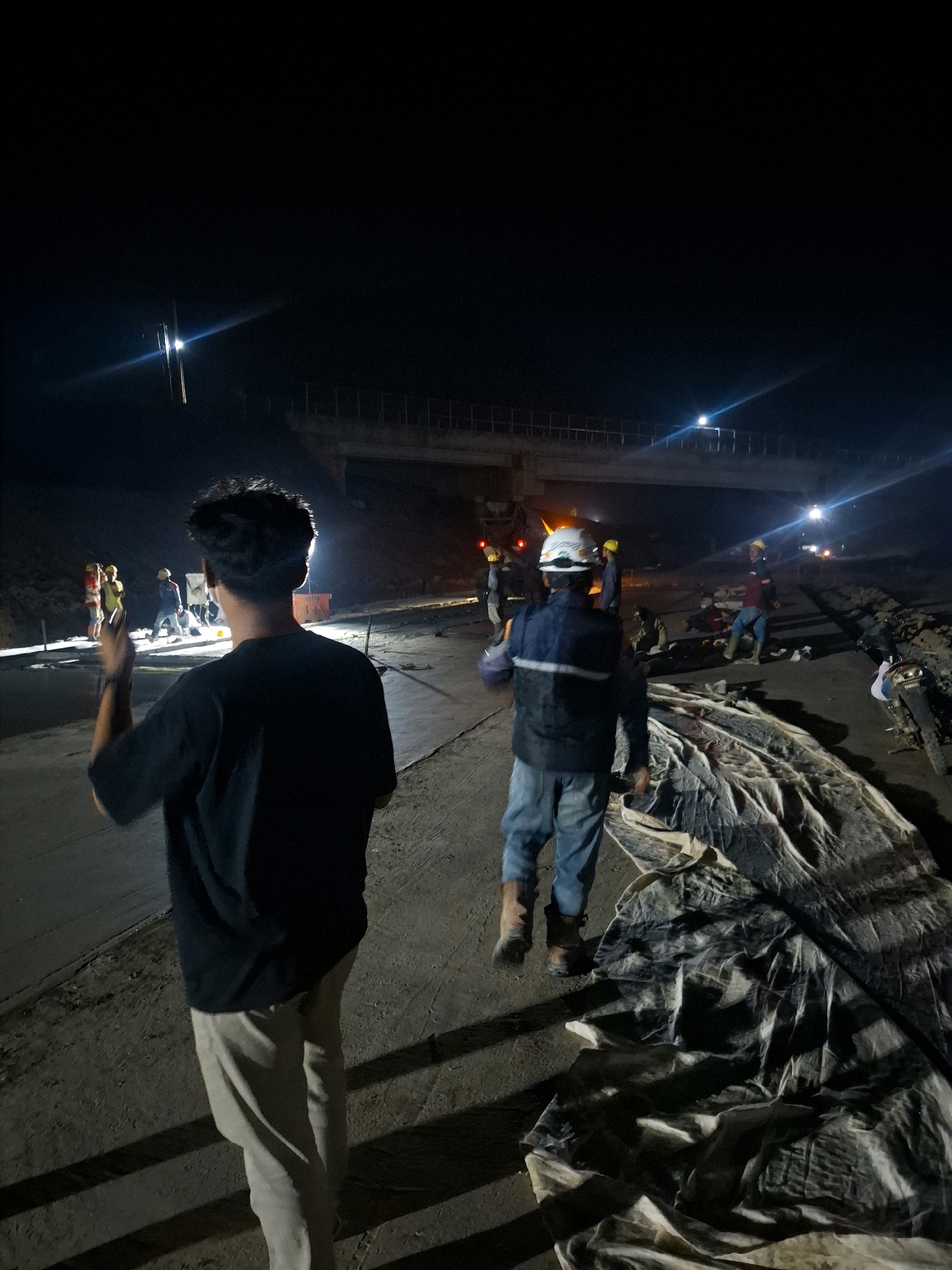 This is a nighttime, outdoor photograph taken at a construction site in Karangtengah, Indonesia. A group of construction workers are present, illuminated by artificial lights. In the foreground, facing away from the camera, a person wearing a black t-shirt and light-colored pants stands with their back to the viewer. Their left arm is raised, with their index finger extended. Behind this individual, and slightly to the right, another person wearing a hard hat, a dark vest over a blue long-sleeved shirt, and blue pants is walking. They are carrying a yellow object.
In the background, several other individuals in hard hats and safety vests are visible, some appearing to be working. There is construction equipment, including what appears to be a truck with flashing lights. A large, light-colored tarp is spread out on the ground to the right of the frame. An elevated structure, possibly a bridge or overpass, is visible in the distance. The environment appears to be a dirt and gravel construction area, with shadows cast by the lighting.
