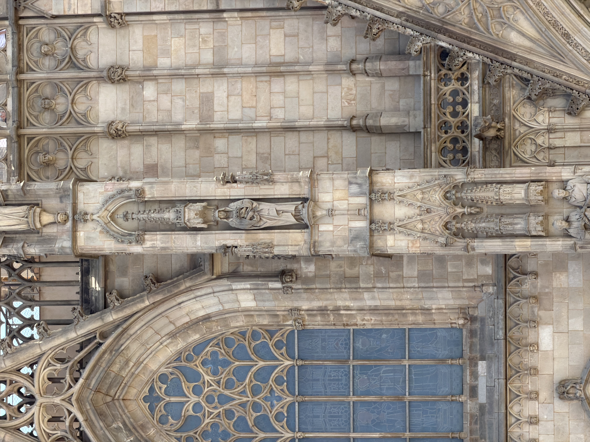 The image shows a close-up, eye-level, exterior view of a Gothic cathedral's facade in Barcelona, Spain. The architecture is characterized by intricate stone carvings, including statuary, gargoyles, and ornate tracery, predominantly in light-colored stone. A large, pointed-arch window with blue stained glass is visible on the left. Niches house statues, with one prominent statue of a woman holding an infant positioned vertically in the center of the frame. Numerous small sculpted faces are embedded in the stonework along a horizontal decorative band. The lighting suggests a daytime setting with sunlight illuminating the facade, casting subtle shadows that highlight the architectural details. The focus is entirely on the building's structure and ornamentation, with no people clearly visible in action.