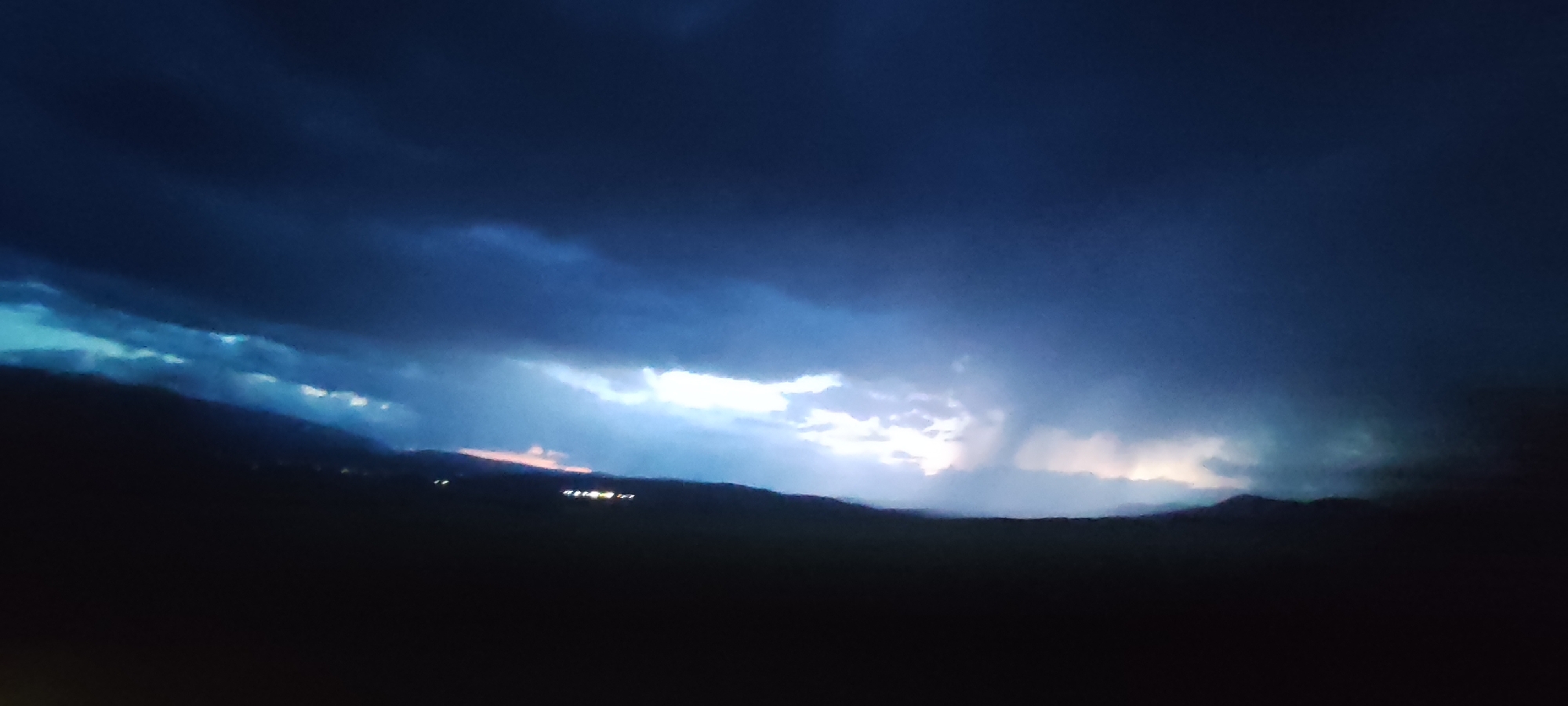 A low-angle, wide shot captures a dark, stormy sky over a silhouetted landscape at dusk or night. The sky is dominated by deep blues and blacks, with a dramatic horizontal band of bright white and pale yellow light breaking through the clouds in the middle distance, suggesting either lightning or the setting sun behind a cloud formation. Below this dramatic sky, a dark, undulating line represents a mountain range or hills. Along the ridge of this silhouette, a scattered line of small, warm-toned lights, likely from buildings or streetlights, are visible. The foreground is entirely black, offering no discernible details. The overall mood is somber and atmospheric, hinting at an approaching storm or the aftermath of one. The location is not identifiable from the image alone.