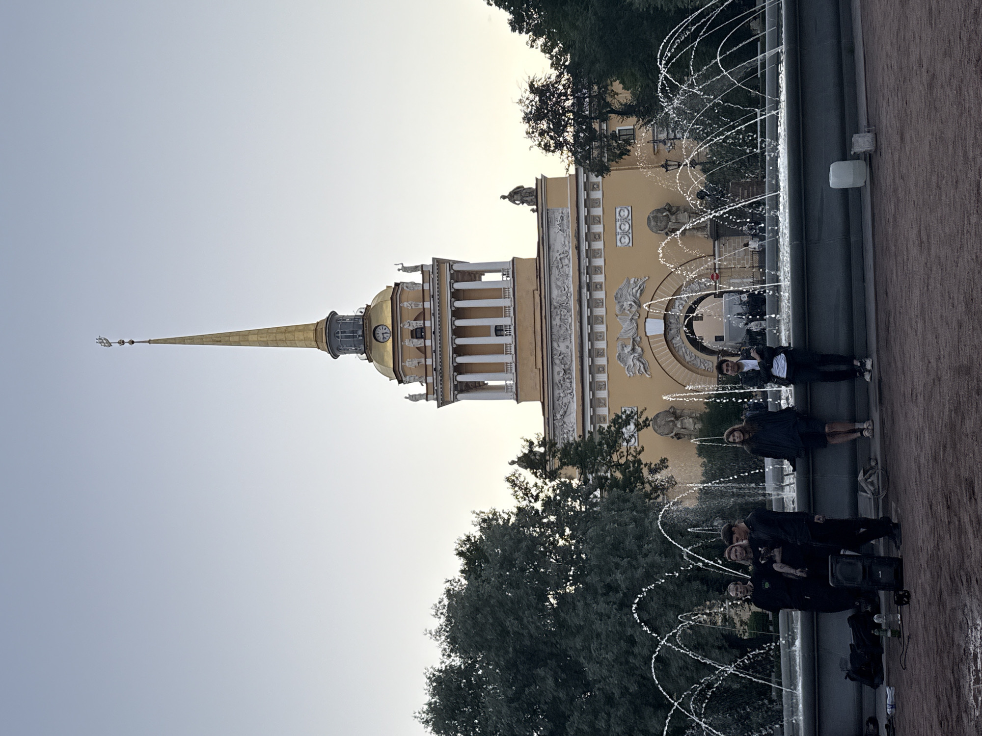 In Saint Petersburg, Russia, in front of the Admiralty Building, a fountain is active. Six people are standing near the fountain. Some are standing in small groups, and one is standing alone. There is a speaker and other equipment on the ground in front of the fountain. The sky is clear and the sun is low.