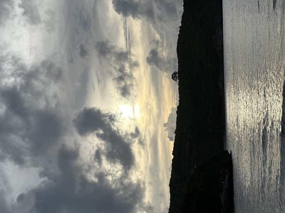 The image displays a coastal landscape in Sekongkang, Indonesia, under a cloudy sky during a period of low sun. The upper half of the frame is dominated by thick, dark grey clouds, with a bright, diffuse sun appearing through a break in the cloud cover, centered horizontally and positioned in the upper middle section. This sunlight casts a yellowish hue on the surrounding clouds. A prominent, dark green landmass, heavily covered in dense vegetation, forms a curved horizon line across the middle of the image. On the highest point of this landmass, slightly right of center, a single, distinct tree with a rounded canopy is silhouetted against the sky. The lower half of the image depicts a body of water, likely the sea, with a gently rippled surface. A bright, elongated reflection of the sun shimmers on the water, extending from the horizon into the foreground. Rocky formations are visible along the waterline at the base of the landmass on the left side. No individuals, animals, or man-made structures are present or engaged in any discernible actions within the scene.