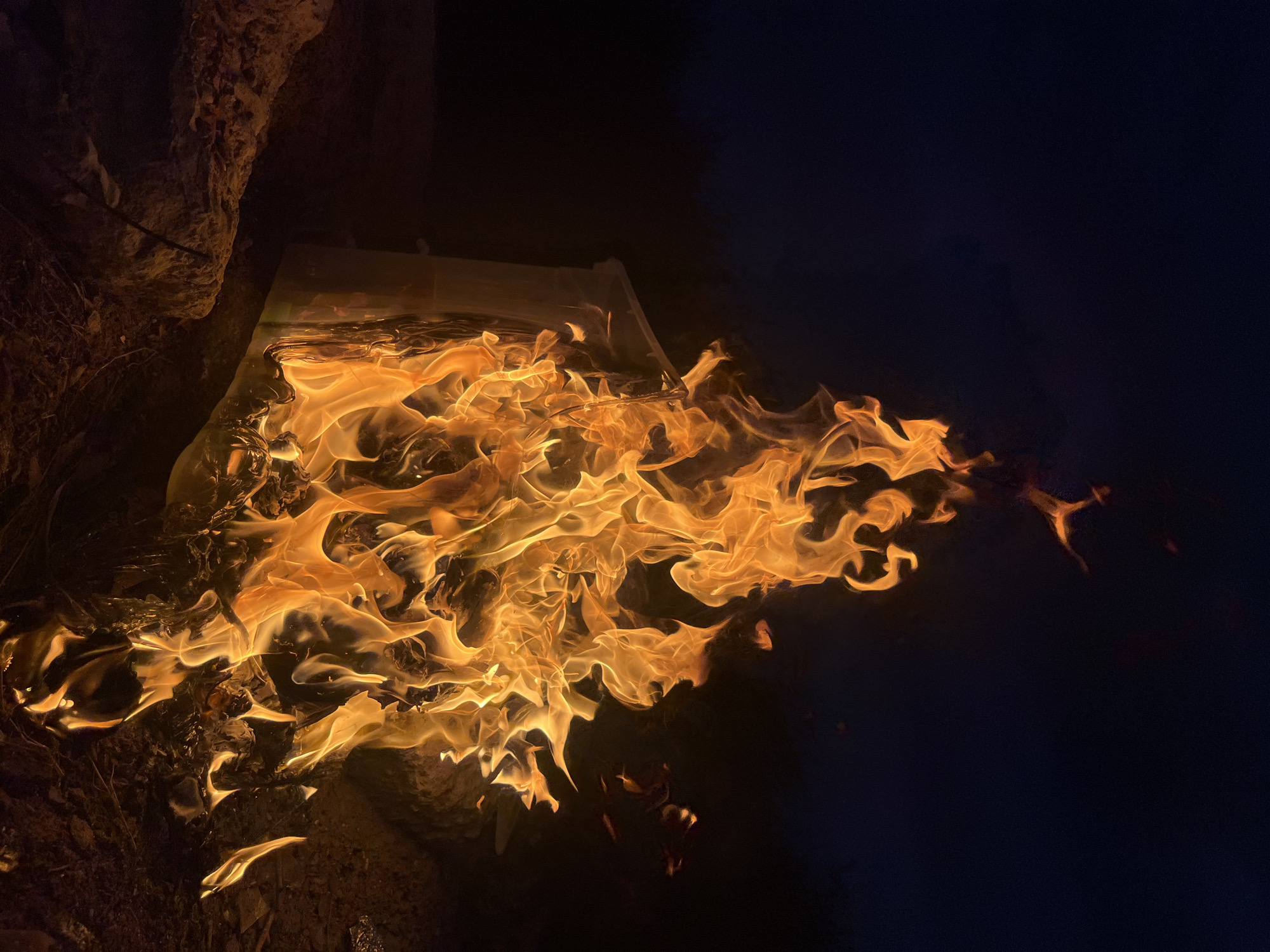A close-up, vertical shot captures a bright orange fire engulfing the side of a white object, possibly a structure or a large piece of debris, set against a dark, night sky. The flames are dynamic and extend upwards, with some smaller embers visible in the background. The foreground shows the ground with rocks and dry vegetation. The scene appears to be outdoors, with no people visible. The location is not specified in the image or any provided metadata.