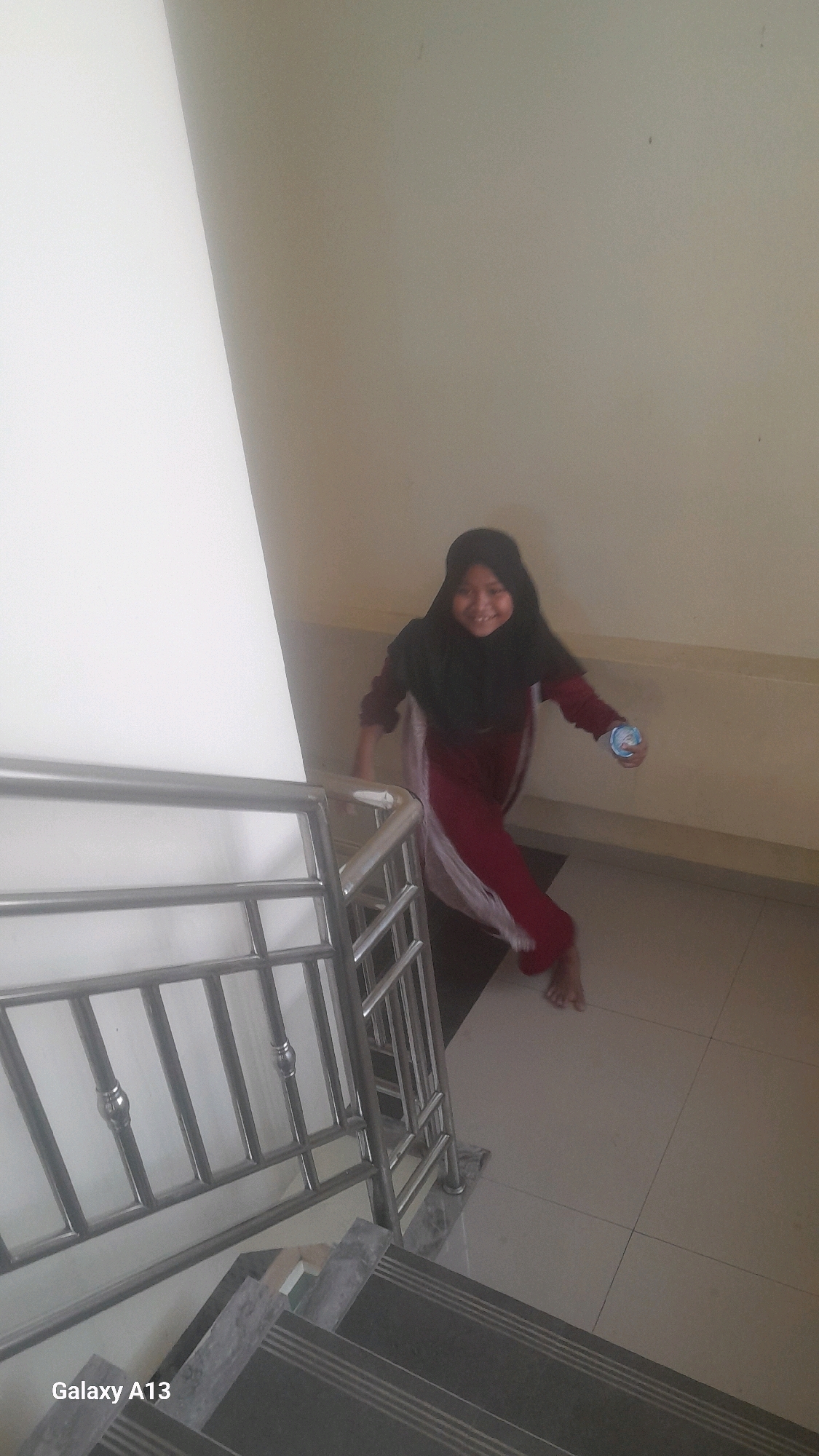 A young girl wearing a hijab and a maroon dress stands on a tiled floor next to a metal railing on a staircase. She is smiling and holding a small cup in her right hand. The staircase is made of dark gray tiles with lighter gray stripes. A white wall is visible to the left of the girl. The scene appears to be indoors.