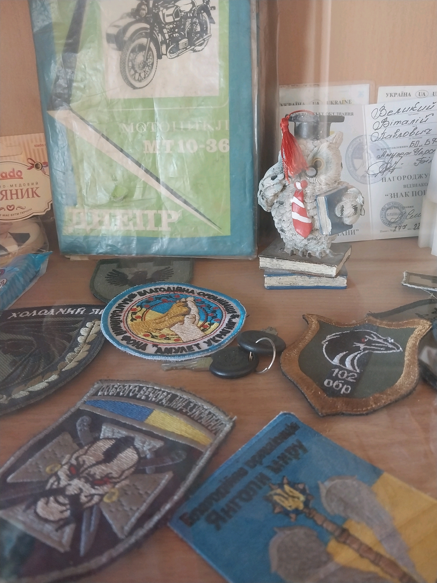 The image is a close-up shot of a collection of items displayed on a wooden surface, possibly a shelf or tabletop. In the background, a large blue binder or folder contains an image of a motorcycle with a sidecar. To the right of this binder, a small figurine of an owl wearing a graduation cap and gown is perched on a stack of books. Several documents, including what appears to be a diploma or award with Ukrainian text, are visible behind the owl.
In the foreground and midground, a variety of patches and small objects are arranged. These include:
* Several circular and shield-shaped embroidered patches, many featuring military or national symbols, including the Ukrainian flag and a trident emblem. One patch has the text "102 обр" (likely indicating a military unit).
* A small packet with text that includes "slado" and "медовий" (honey).
* A keychain with two dark-colored keys.
* A blue and yellow rectangular patch with an image of wings and a mace, also with Ukrainian text.
The lighting suggests the items are indoors, possibly in a display case or on a shelf in a private collection or museum. There are no people or animals visible in the image. The overall impression is that of a curated display of memorabilia, with a focus on Ukrainian national identity and possibly military history. The specific location in Dnipro, Ukraine, cannot be determined from the visual information provided.