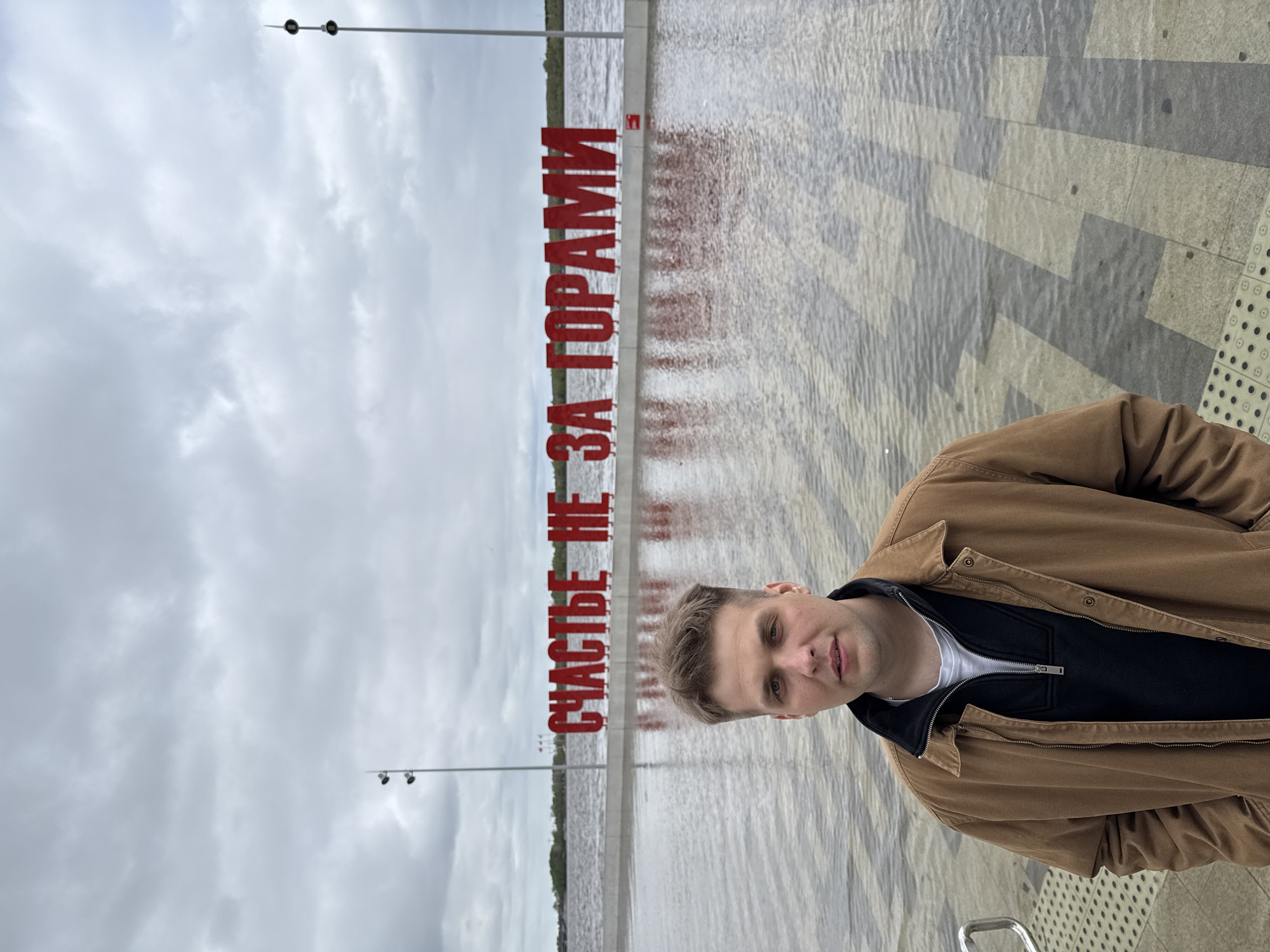 A man stands on a patterned gray pavement next to a body of water. Behind him, large red Cyrillic letters spell out a phrase. The sky is overcast. This appears to be a photograph taken in Perm, Russia. The red letters are part of a public art installation near the Kama River. The man is wearing a brown jacket.