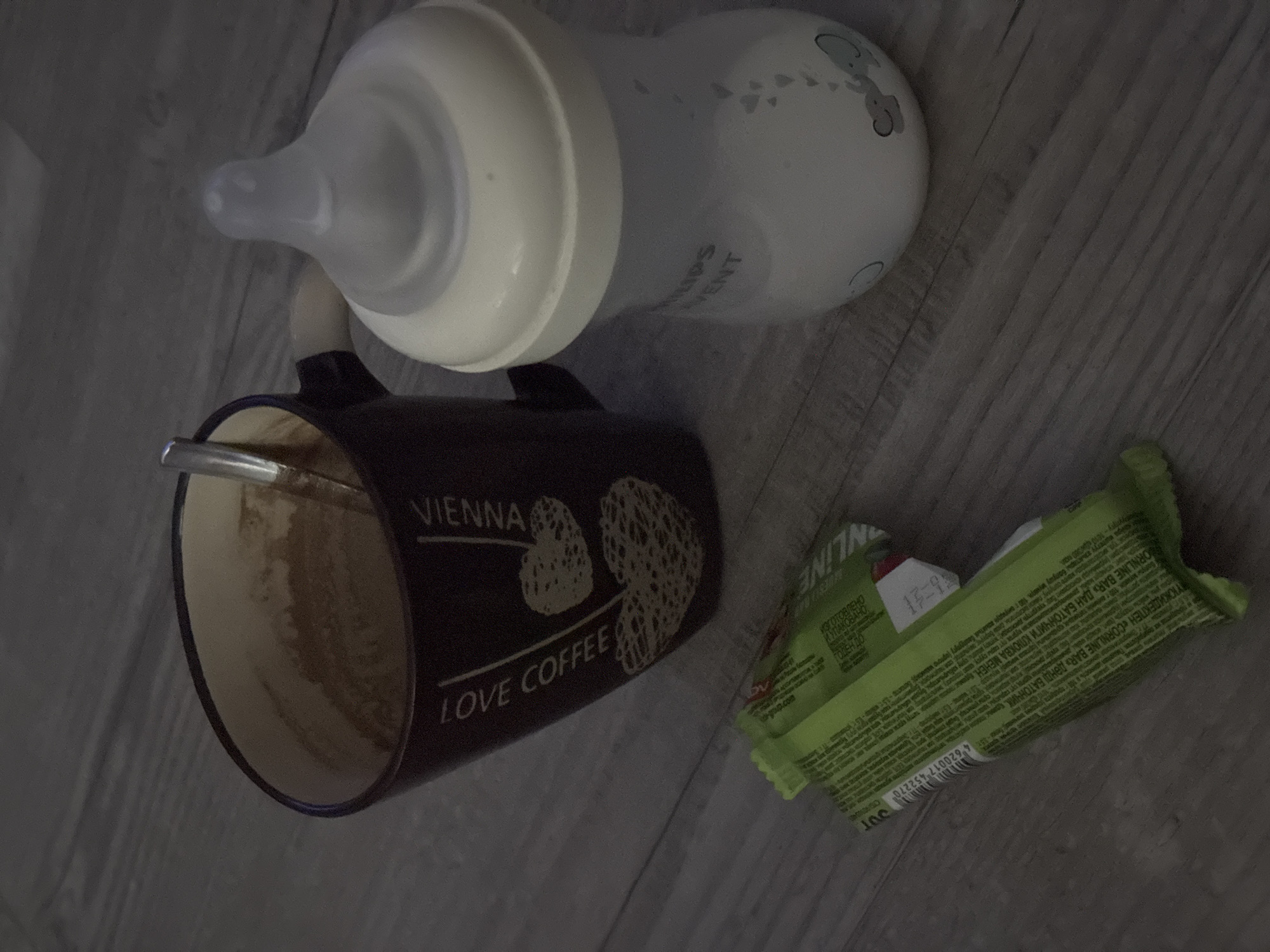 An indoor, eye-level, medium shot depicts a wooden floor with three objects on it. In the upper left is a dark brown mug with a spoon inside and a coffee residue. The mug has text that reads "LOVE COFFEE VIENNA" and heart designs. To the right of the mug is a baby bottle with a white nipple and a partially filled white liquid. The bottle has "PHILIPS AVENT" printed on it. In the lower left are two green candy bars. The lighting is dim.
