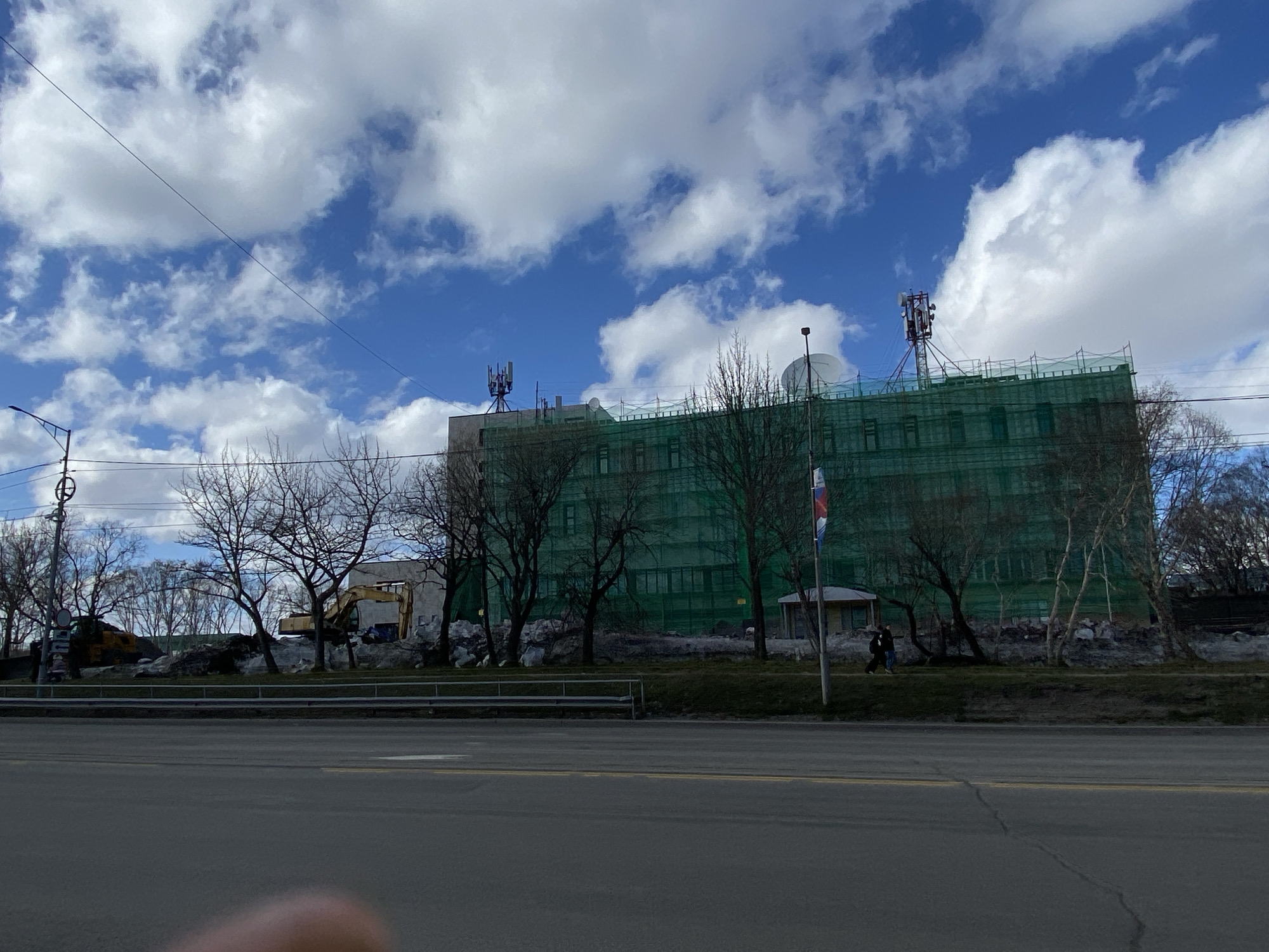 The image shows a street scene in Petropavlovsk-Kamchatsky, Russia. In the background, a large building is covered in green netting. Two communication towers are visible on the roof of the building, as well as a satellite dish. In front of the building are several bare trees. A Russian flag is waving near a small structure. An excavator and another piece of construction equipment are visible among piles of rubble near the trees. A person is walking on the sidewalk along the edge of the road. The foreground is dominated by an asphalt road. Above, the sky is blue with scattered white clouds.