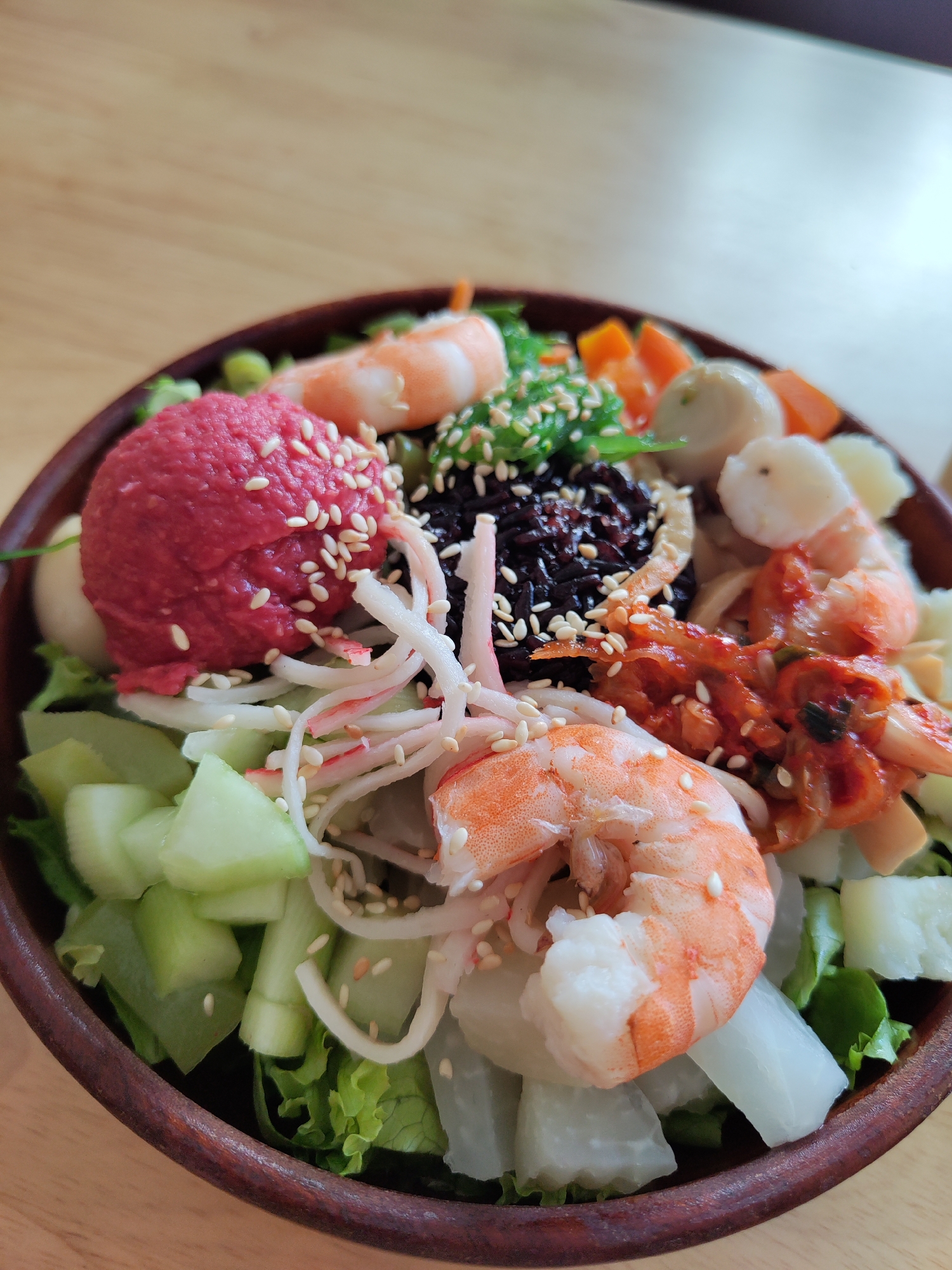 A close-up, high-angle shot depicts a bowl of poke-style food. The bowl appears to be made of dark wood.
Ingredients include cooked shrimp, shredded imitation crab meat, diced cucumbers and white radish, lettuce, black rice, kimchi, and a scoop of what appears to be beet puree, covered with sesame seeds. Other ingredients include green onions, carrots and some types of small white balls.
The bowl sits on a light brown wooden surface, with bright indoor lighting reflecting off the surface and the bowl's contents. The image was taken in Da Lat, Vietnam.