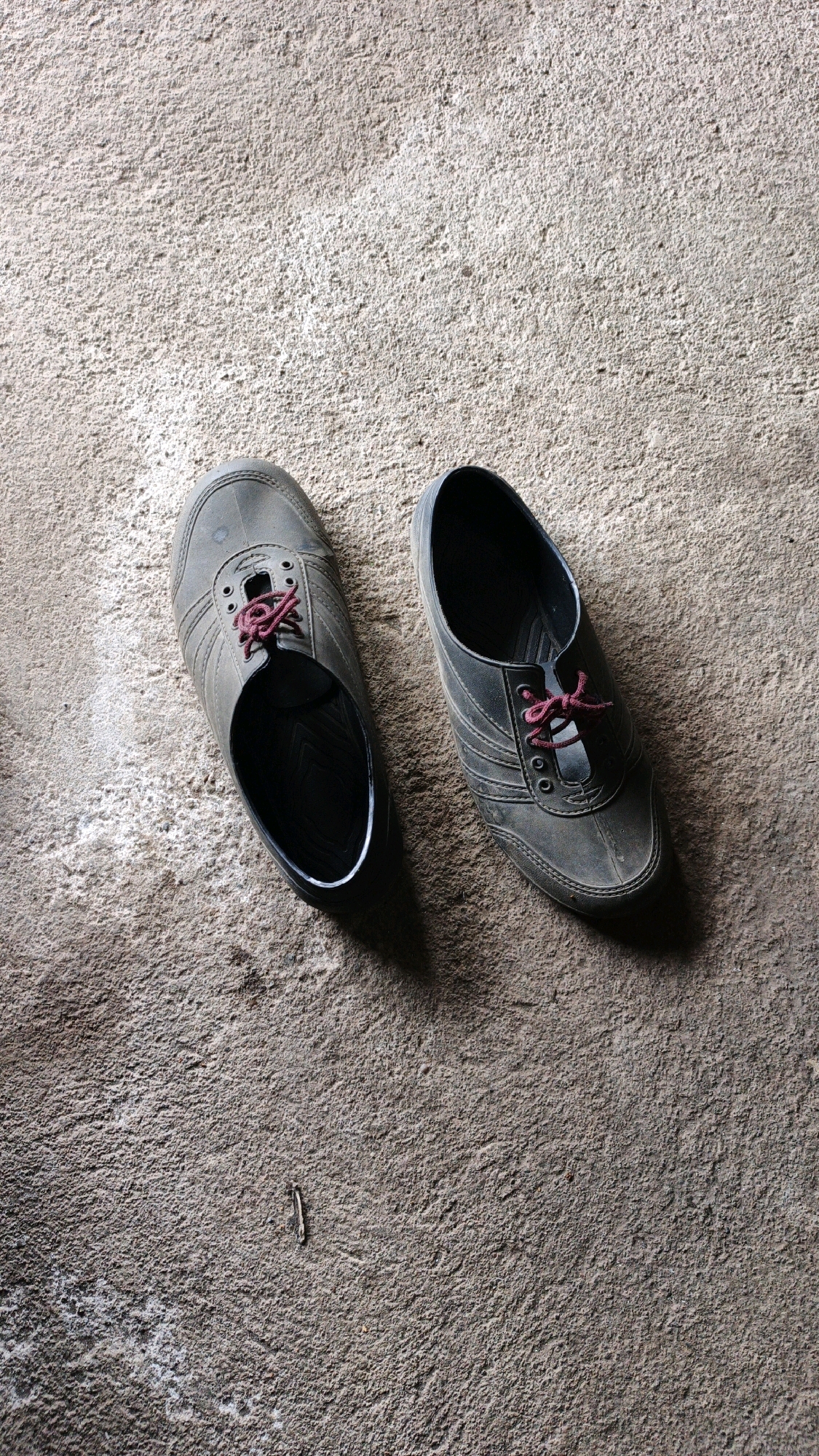 Two dark gray rubber shoes, each with dark pink laces, are placed on a rough, gray concrete surface. The shoes are oriented with their toes pointing away from the viewer and slightly to the right and left, respectively. The left shoe is slightly more to the left of the frame, and the right shoe is slightly more to the right. Shadows are cast to the left of each shoe, suggesting a light source from the upper right. The concrete surface appears somewhat worn and textured. The image is taken from a high-angle, overhead perspective. The scene is devoid of human presence or activity, with only the shoes and the concrete floor visible. There is no indication of the specific location within Ngranti, Indonesia, beyond it being a general outdoor or semi-outdoor environment with a concrete floor.