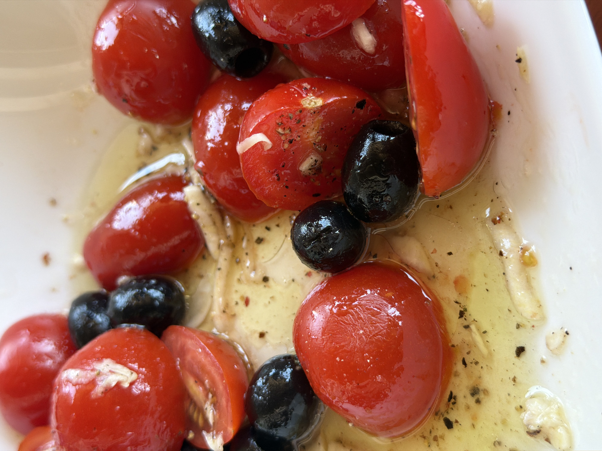 A close-up, top-down view reveals a partially consumed salad within a white dish. The salad consists primarily of glistening, bright red cherry tomatoes and smaller, round black olives, all coated in an oily dressing. Some tomatoes are whole, while others have been halved or sliced. Thin, white strands, possibly garlic or cheese, are scattered throughout the dish. The dressing appears to be a light yellow or golden color, speckled with black pepper. The composition suggests a simple, yet flavorful, Mediterranean-style dish. There is no indication of the location being Budva, Montenegro.