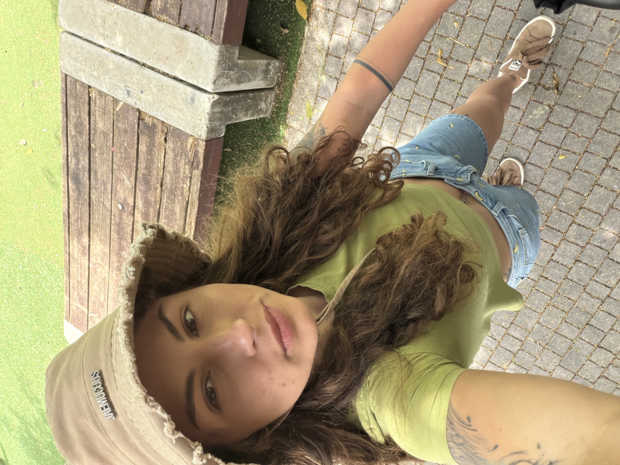 A woman stands on a paved area composed of square paving stones. She is wearing a tan bucket hat with a frayed edge, a light green short-sleeved shirt, and denim shorts with a pattern of small yellow objects. Her shoes are tan suede. She holds a device in her extended right arm, taking a selfie. She has brown hair and visible tattoos on her arms. To the left of the frame is a wooden structure with concrete supports. A small patch of green ground cover is visible. The scene is likely in Kiryat Bialik, Israel, but there is no direct evidence in the image to confirm this.