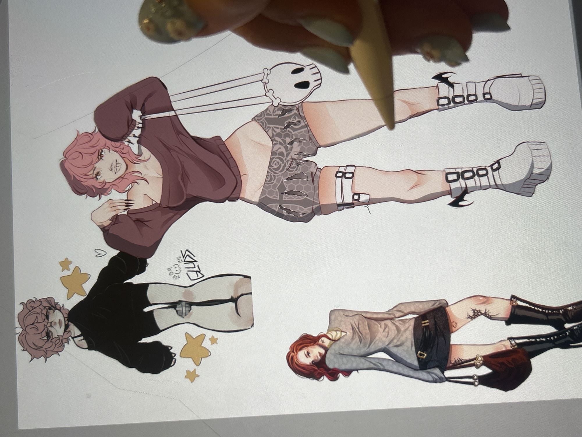 The image displays a digital illustration featuring three anime-style characters. The foreground shows a partial view of a hand with elaborate nail art, holding a stylus, which is interacting with the digital artwork.
The first character, depicted at the top left, is a pale-skinned figure with messy pink hair and round glasses. They wear an oversized black off-the-shoulder sweater and black shorts, with stylized bandages on their legs. Yellow stars and a heart are drawn around this character.
To the right is a second, taller character with similar messy pink hair and visible fangs. They are wearing a maroon off-the-shoulder sweater and patterned shorts, with white platform boots featuring black buckles and bat-wing accents. A white skull-shaped purse hangs from their shoulder.
At the bottom left, a third character with long, wavy red hair is shown. She wears a light grey long-sleeved top with a cowl neck and a short black skirt. She is also wearing black heeled boots and carrying a dark red handbag. Tattoos are visible on her legs.
The background is a plain white digital canvas. The image does not contain any elements related to Cupra Marittima, Italy.