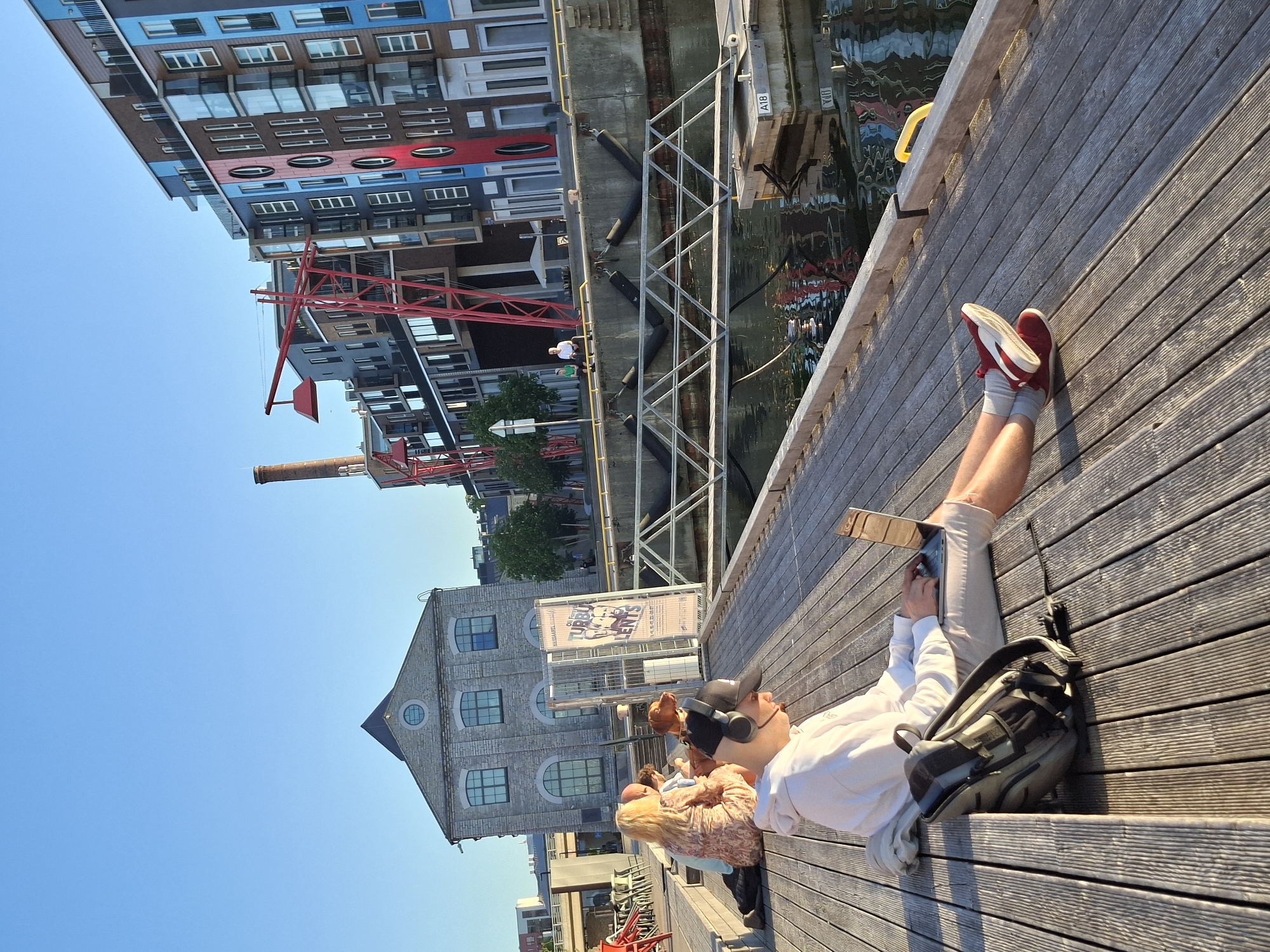 The image captures a sunlit waterfront scene in the Noblessner district of Tallinn, Estonia, characterized by a blend of repurposed industrial architecture and modern residential buildings.
In the immediate foreground, a young man is seated on a light-colored wooden plank deck. He wears a white hoodie, grey shorts, a black baseball cap, and black over-ear headphones with an attached microphone. He is actively using a silver laptop, which rests on his lap. A black and grey backpack is positioned to his left on the deck. His legs are extended, wearing white socks and red sneakers with white soles.
Further back on the left, a group of three individuals and a brown dog with a collar are seated on a slightly elevated section of the wooden deck, appearing relaxed. One woman with blonde hair wears a patterned dress.
Across a calm body of water, a concrete retaining wall with black rubber fenders is visible. A metal truss bridge structure extends over the water. On the far bank, two people are seated on the concrete wall near a white patio umbrella. A floating dock, marked "A18," is partially visible in the water.
The background features prominent architectural elements. To the left, a substantial, historic-looking grey stone building with multiple arched windows and a prominent circular window on its facade stands under a dark, pointed roof. To the right of this building, a tall vertical banner with Estonian text is displayed. Behind the waterfront, a series of modern, multi-story apartment buildings line the street. These buildings showcase varied facades, including sections with brown, blue, and red paneling, balconies, and a mix of rectangular and circular windows. A tall, reddish-brown brick industrial chimney rises behind these structures, alongside large, red, abstract crane-like art installations or structural components.
The sky is clear and bright blue, and the scene is illuminated by strong sunlight, casting long, distinct shadows on the wooden deck and across the water. The water's surface reflects the surrounding buildings and the sky.