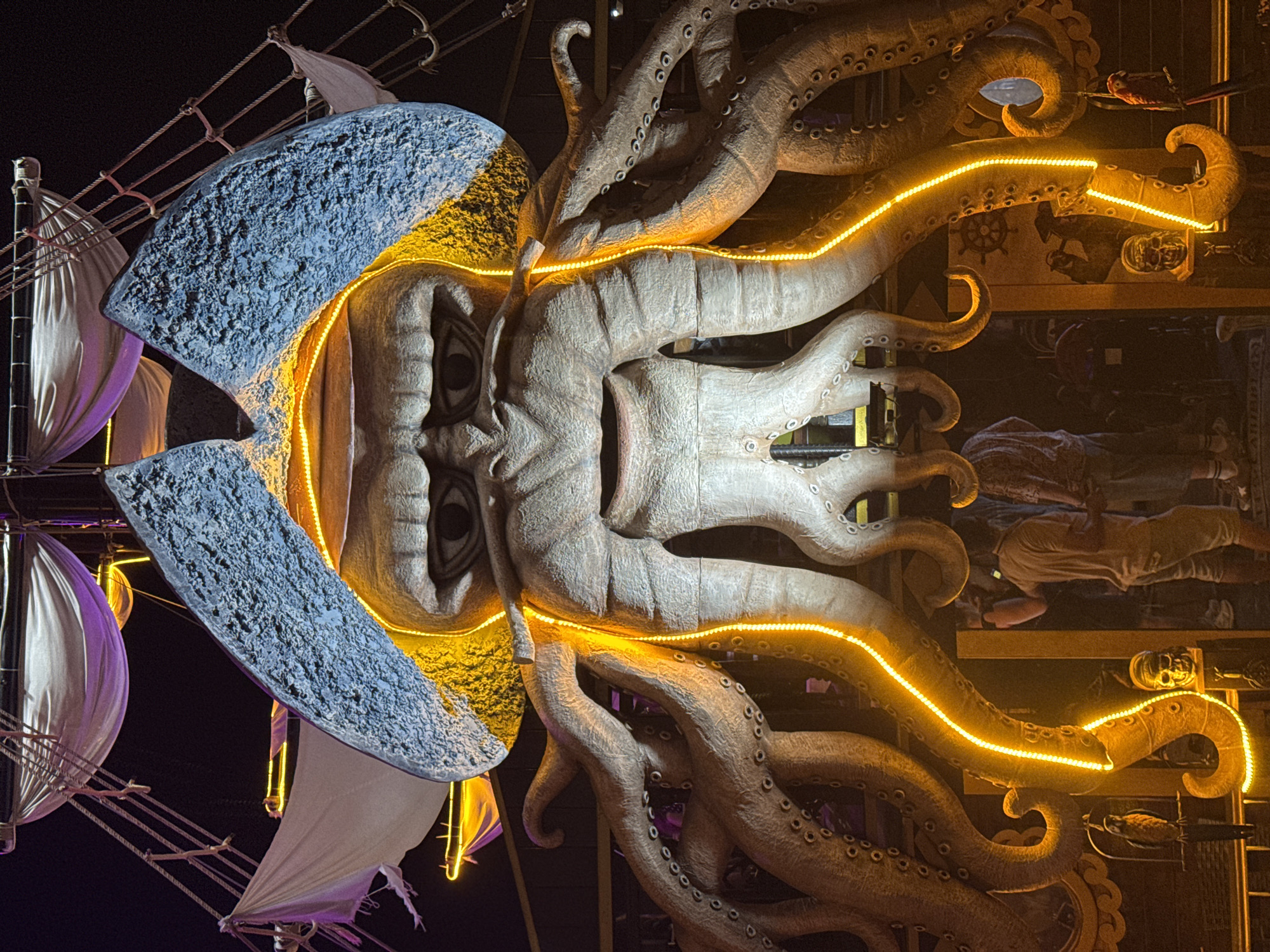This is a nighttime, eye-level, medium shot of a large, theatrical prop resembling a Kraken with a pirate hat. The prop dominates the frame, with its large, textured tentacles arching downwards and illuminated by orange LED strip lights. The tentacles are studded with suction cups. Above the tentacles is a massive, stern face with bulging eyes and a gaping mouth, wearing a giant, textured blue pirate hat with a gold brim. In the background, parts of a ship's rigging and sails are visible, also illuminated with purple and white lights against a dark sky. Below the Kraken, a few people are visible standing in what appears to be an entrance or walkway. To the left and right of the entrance are decorative elements, including a skull and a perched parrot on the left, and a ship's wheel and another skull on the right. The overall scene suggests a themed attraction, possibly at a fairground or entertainment venue. The location is identified as Batumi, Georgia.