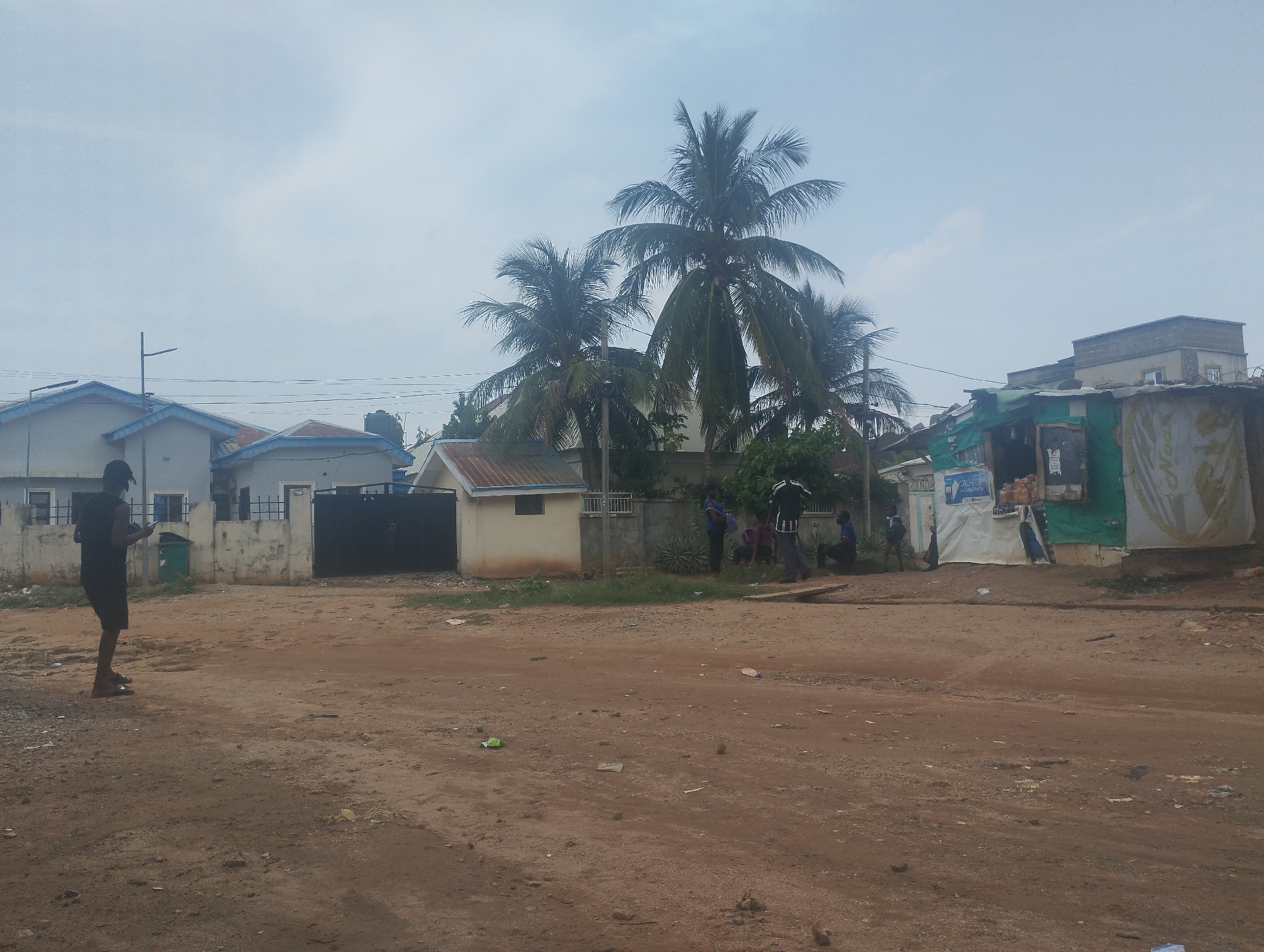 The image depicts a street scene in Gwarinpa, Nigeria. A man stands on the left side of a dirt road, looking at his phone. Across the road, there are several buildings, a black gate, and palm trees. A group of people are gathered near one of the buildings. The building on the right is a makeshift structure with tarp coverings. The sky is overcast. There is trash scattered across the ground.