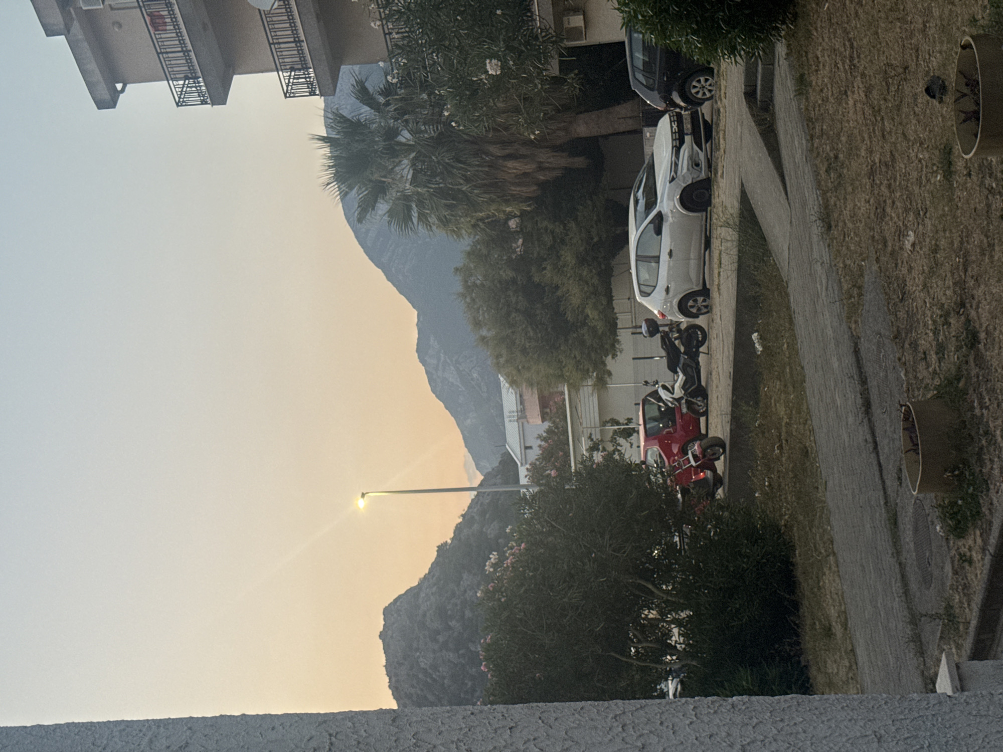 The image shows a street scene in Sutomore, Montenegro, during dusk. A residential building with balconies is visible on the right. In the foreground, a paved walkway leads to a ramp, with dry grass and planters on either side. Several vehicles are parked along the street, including a red car, a white hatchback, and a black car. Two motorcycles are also present. Lush green trees and bushes, some with pink flowers, are scattered throughout the scene. In the background, mountainous terrain and buildings are silhouetted against a pale orange sky, indicating the setting sun. A streetlamp illuminates the area.