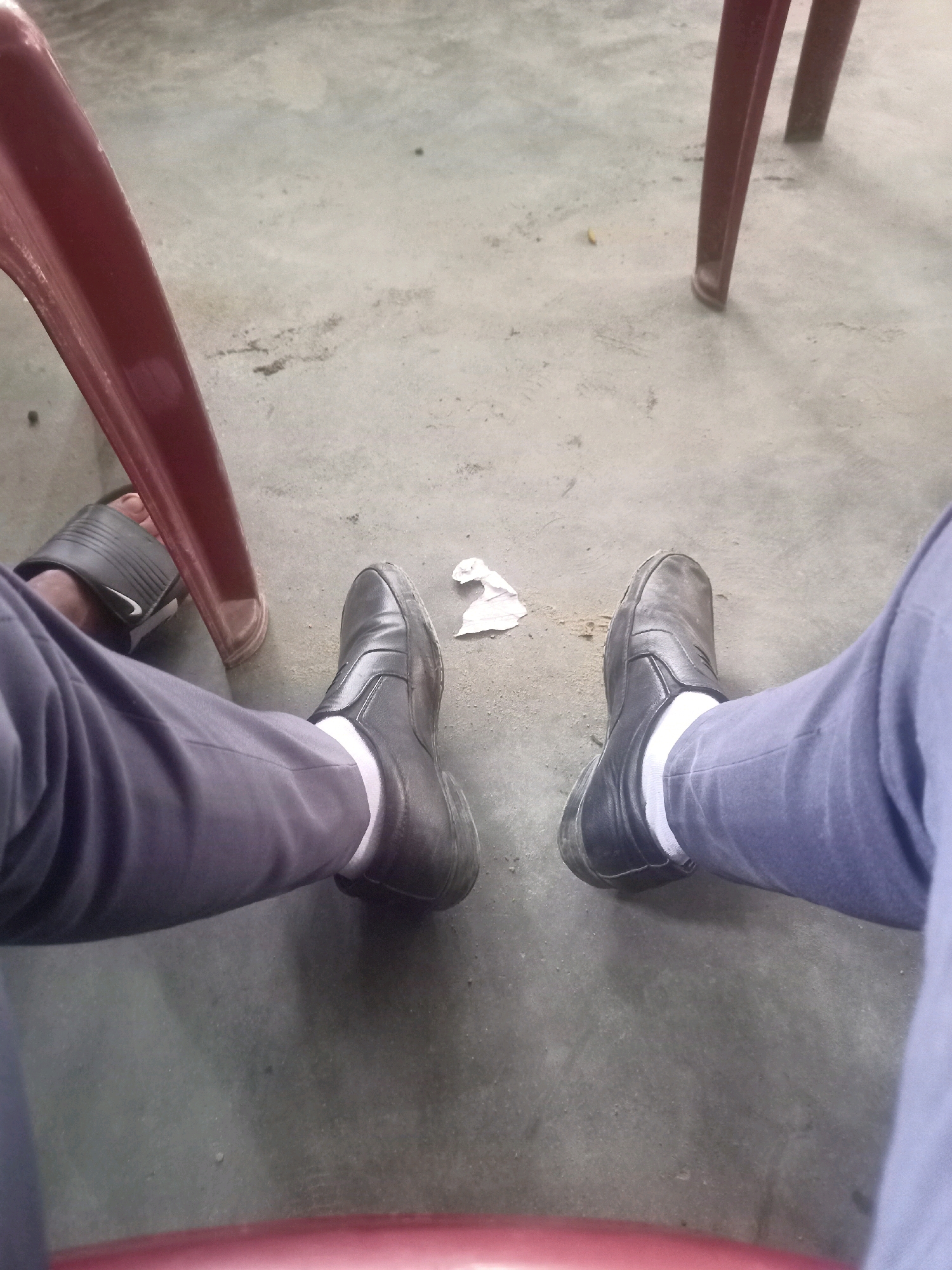 The image captures a first-person perspective of someone sitting on a concrete floor, likely in Hatsinigmari, India. Two legs, clad in dark gray trousers and white socks, are visible. One foot is adorned with a black slip-on shoe, while the other is wearing a black Nike sandal. A crumpled piece of white paper rests on the floor between the feet. In the background, the legs of a red plastic chair are partially visible. The floor appears to be a rough, textured concrete surface, with some scattered debris. The lighting suggests an indoor setting with ambient light.