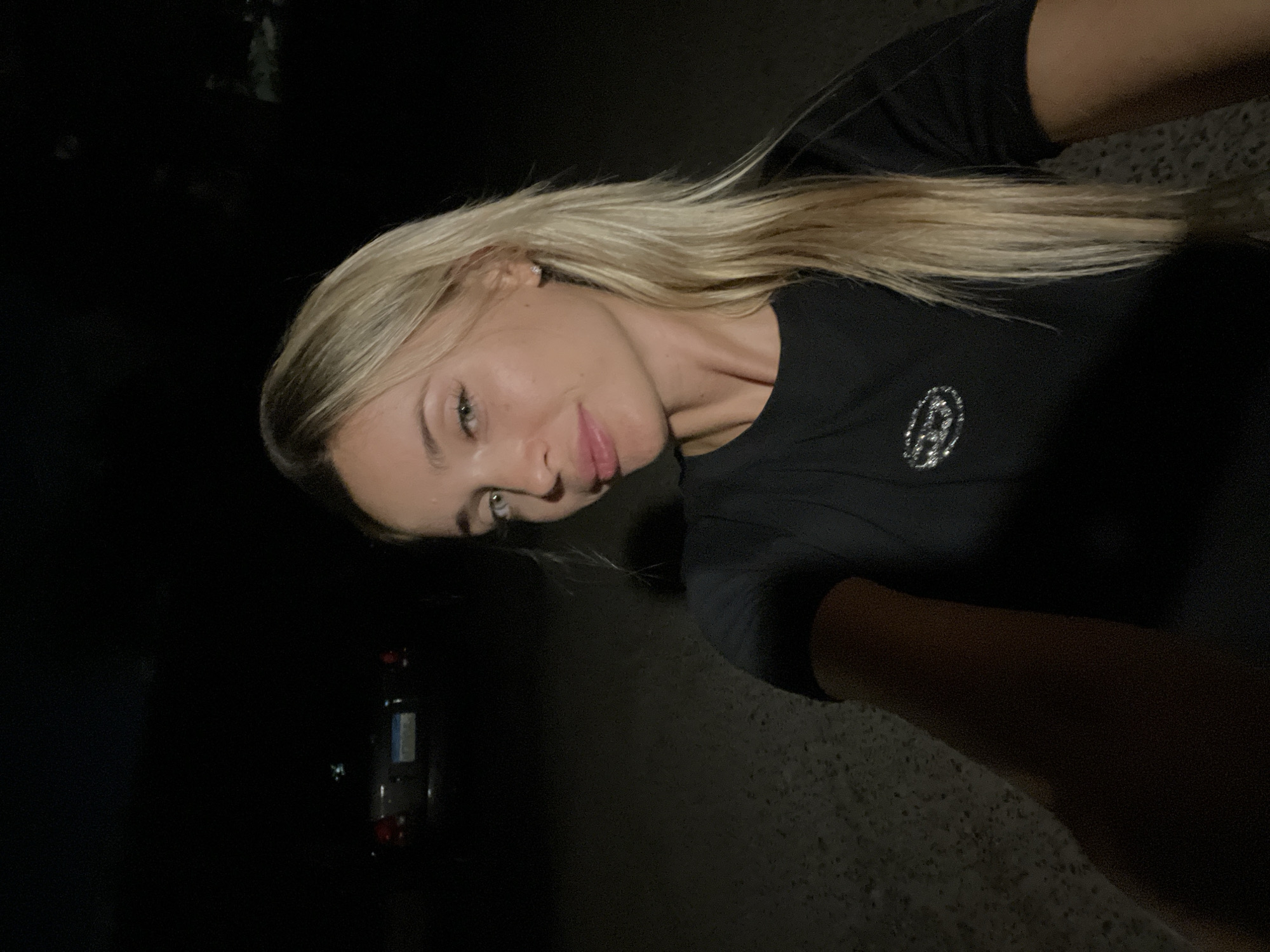A blonde woman with light skin and green eyes takes a selfie at night. She is wearing a black t-shirt with a circular logo on the left chest. Her right arm is extended towards the camera, with her hand out of frame. In the background, a dark car is parked on a gravelly surface, with its taillights dimly illuminated. The overall environment appears to be outdoors, likely in an unlit area of Sharm El Sheikh, Egypt, given the presence of a car and the nighttime setting.