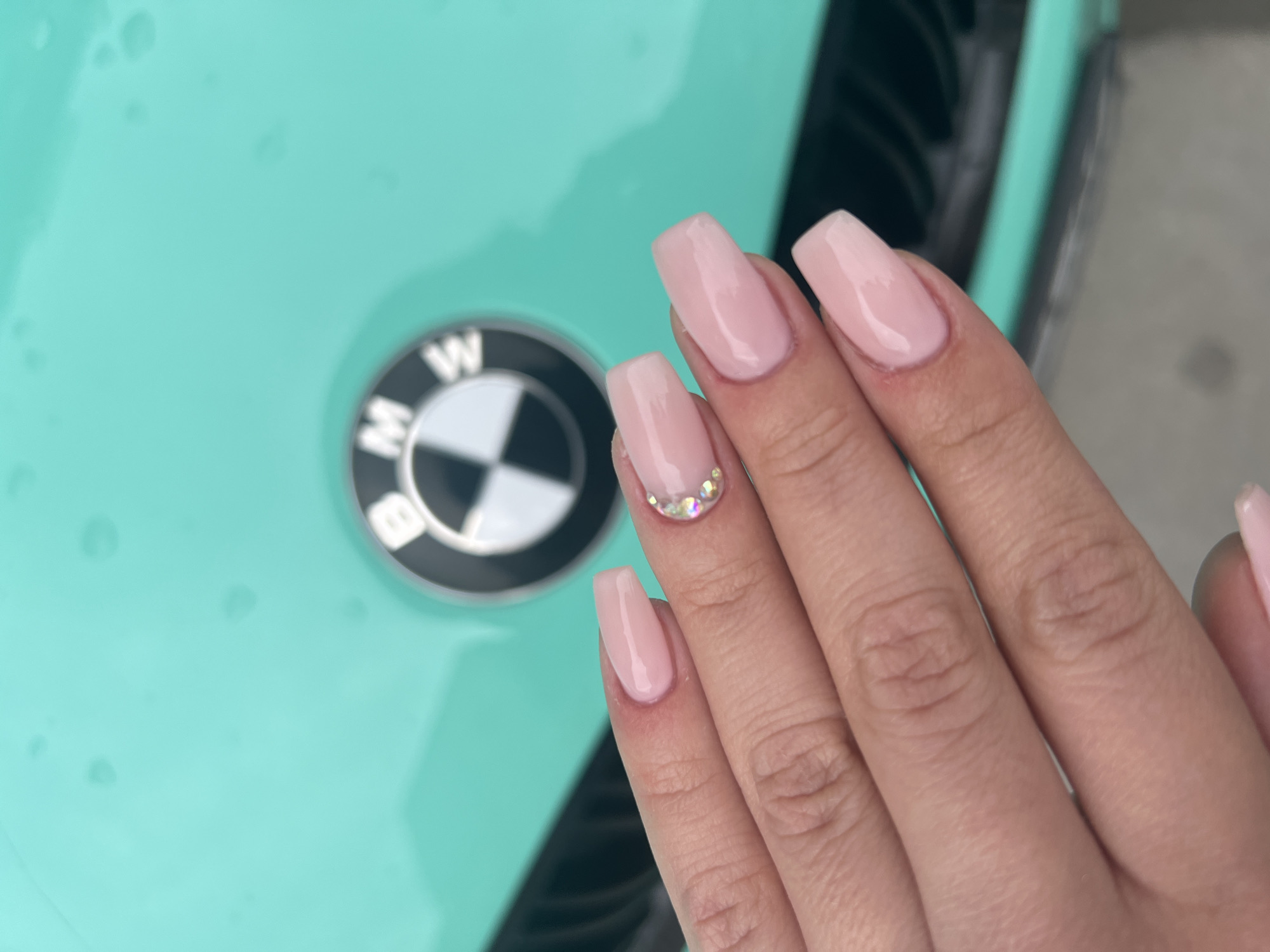 A close-up shot shows a hand with pink acrylic nails positioned near a light green BMW car hood with the BMW logo visible. One nail is decorated with small, iridescent crystals. The location is not evident from this image.
