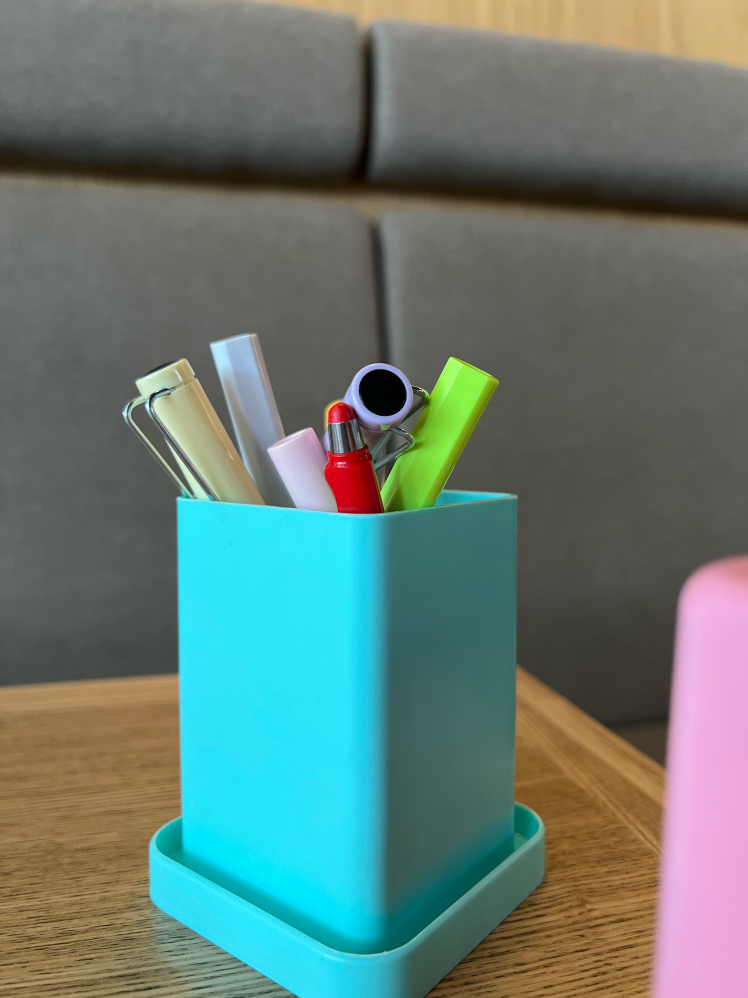The image shows a close-up of a light blue pen holder filled with several writing utensils and a small red lipstick on a wooden surface. The pen holder contains a light yellow pen, a white pen, a light pink pen, a red lipstick, a purple object with a black circle, and a bright green marker. In the background, a grey upholstered couch is visible. The scene is indoors and appears to be on a table. There is a hint of a pink object to the right of the pen holder, out of focus. The image does not provide any information about the location in Krasnoyarsk, Russia.