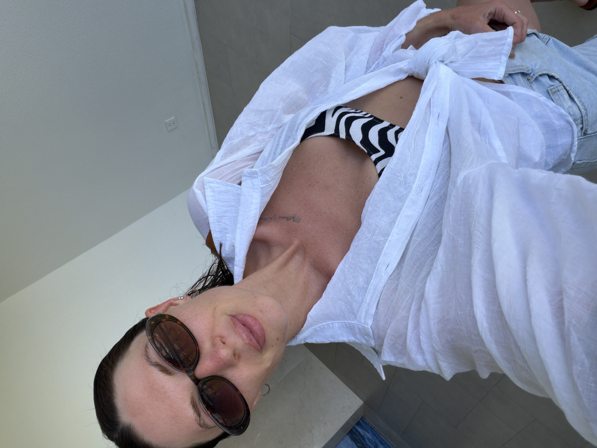 A woman takes a selfie outdoors, wearing sunglasses, a white open button-up shirt over a black and white zebra-print bikini top, and denim shorts. Her hair is dark and wet, and she is wearing hoop earrings and a ring on her right hand. The background shows a light-colored wall and tiled floor, with a glimpse of blue water and a dark structure on the far left. A power outlet is visible on the wall. The scene is a generic outdoor setting with no specific identifying features of Dong Ninh Hoa Ward, Vietnam.