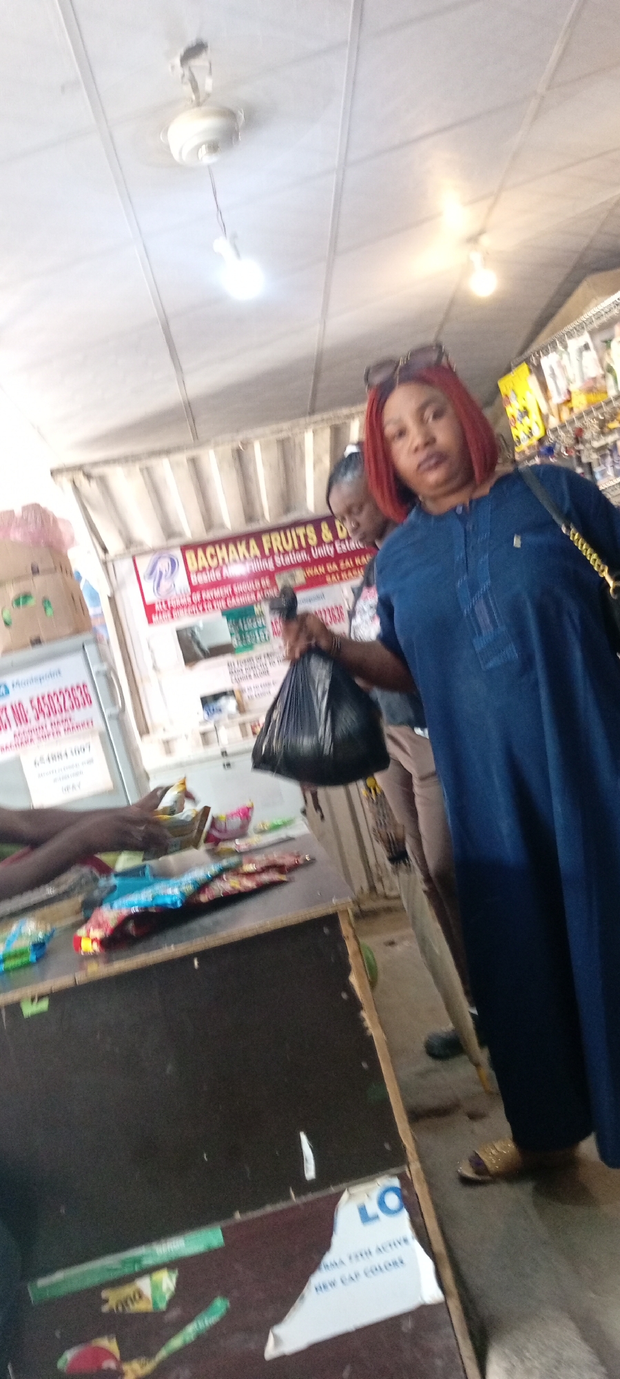 In a market setting, a woman with reddish-brown hair is the central subject. She is wearing a dark blue, long-sleeved, collared garment and is holding a black plastic bag in her right hand. Sunglasses are perched on her head. Behind her, another person, likely a woman, is partially visible. The scene appears to be indoors, possibly a market stall, with a ceiling fan and light fixture visible overhead.
To the left of the main subject, a sign is partially visible, reading "BACHAKA FRUITS & DE" and other details including "Unity Estate." Another sign on a white background displays "CT NO: 5450323636" and "ACCOUNT NAME: BACHAKA SUPER MARKET." On a counter in the foreground, various items, possibly snacks or packaged goods, are displayed. Torn paper with text, including "LO" and "AMA 72H ACTIVE HEW CAP COLORE," is affixed to the front of the counter.
The lighting suggests it could be daytime, given the bright illumination. The overall impression is a bustling market environment, with the woman appearing to be a shopper or vendor. The available text provides specific details about a business named "BASHAKA FRUITS & DE" and a payment reference number.