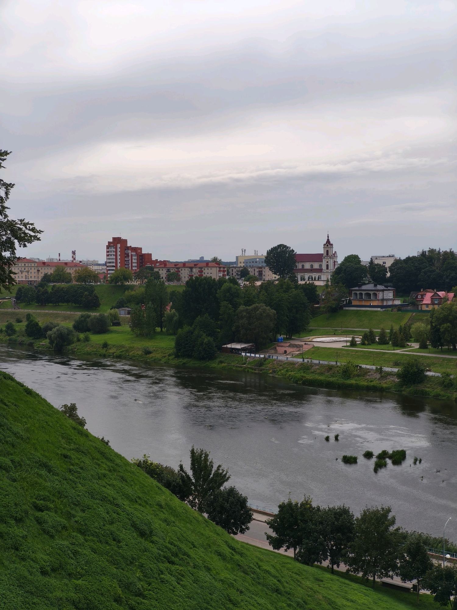 The image shows a panoramic view of the Neman River flowing through the city of Grodno, Belarus. The river is wide and calm, with several small islands of vegetation in the middle. On the right bank, there is a lush green park with trees, walkways, and several buildings, including a prominent white church with a red roof and a tall spire. Further in the background, more buildings, including apartment complexes and industrial structures, are visible. The left bank of the river features a steep, grassy slope leading up to a walkway and more trees. The sky is overcast, with soft, diffused light. The scene depicts a tranquil urban landscape with natural elements.