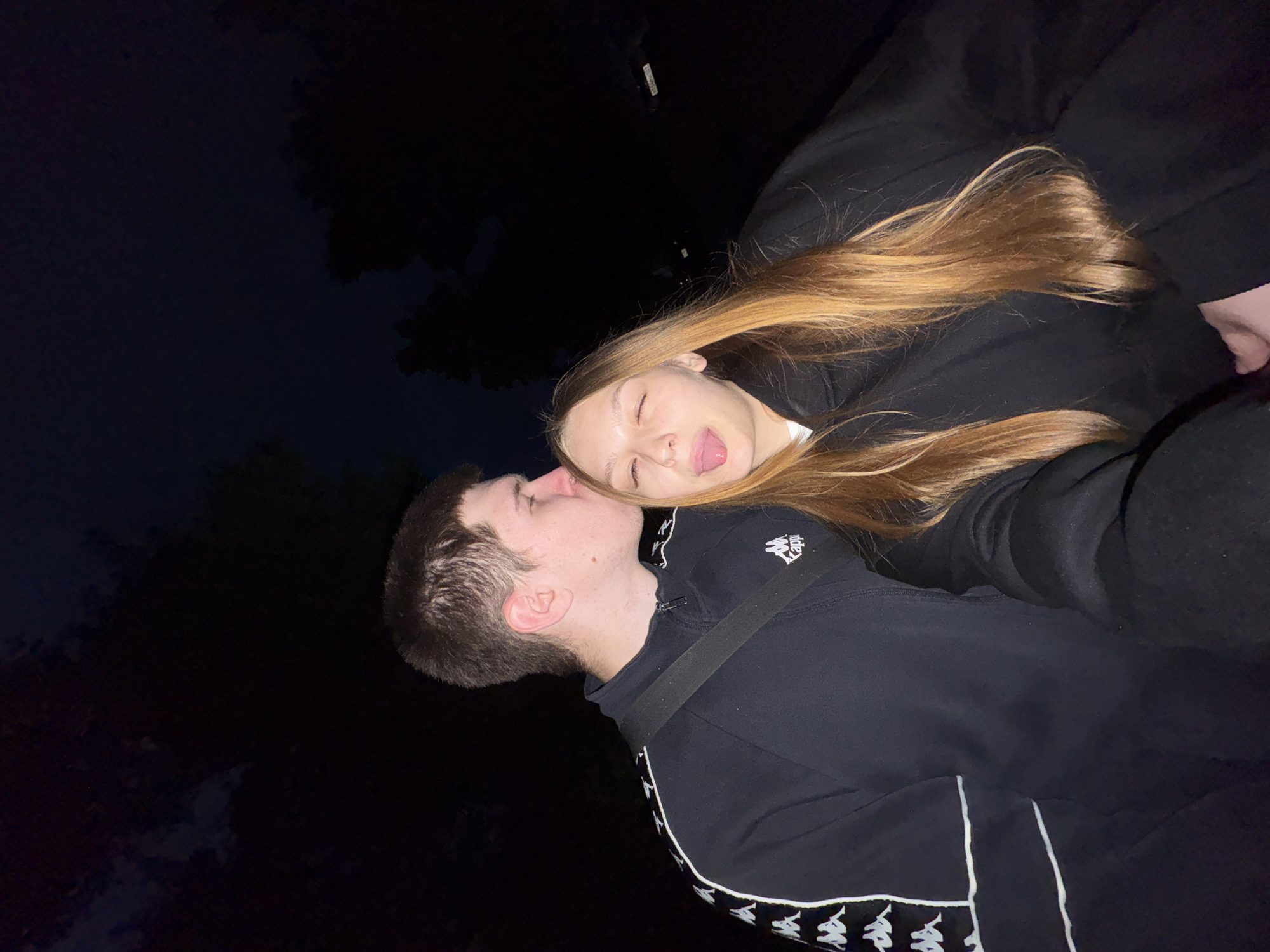 The image depicts two young individuals, a male and a female, in an outdoor setting at night. The male, positioned on the left, has short dark hair and is wearing a black Kappa tracksuit jacket with white trim and logos, and a black strap across his chest. He is leaning in, with his eyes closed, and is kissing the female's forehead or temple. The female, positioned on the right, has long, light brown hair and is wearing a black hooded top. She is looking towards the camera, winking her right eye, and sticking out her tongue in a playful manner. Her left hand is visible at the bottom right, suggesting she is holding the device used to capture the image. The background is dark, with indistinct silhouettes of trees visible against a very dark sky, indicating a nighttime setting. The subjects are illuminated by a direct light source, likely a flash, while the background remains underexposed. No specific environmental details are discernible to identify the location as Chaikovsky, Russia.