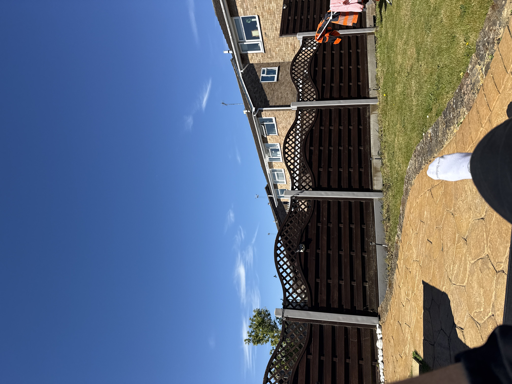 The photograph depicts a suburban scene in Colchester, United Kingdom. A clear blue sky with wispy cirrus clouds dominates the left side of the frame. On the right, a residential property is visible, featuring a two-story brick building with white-framed windows. A dark brown wooden fence with a decorative lattice top runs along the property line, separating it from a green lawn. Clothes hang on the fence. The shadow of a person wearing a white sock is visible in the bottom right corner. The ground is covered with textured, yellow paving stones.