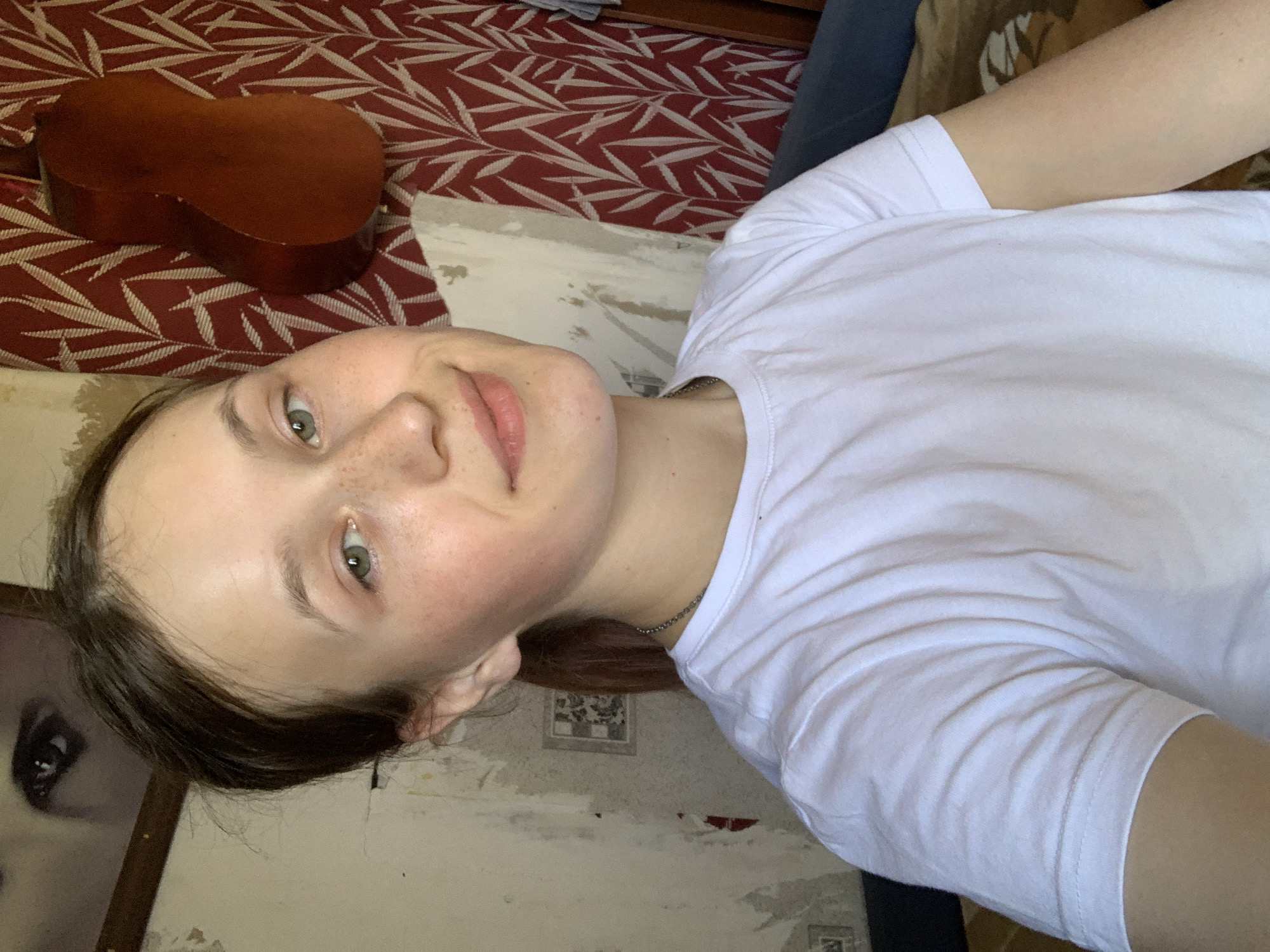 A photograph taken at an unconventional angle depicts a fair-skinned person with light green eyes and freckles, wearing a white t-shirt. The person is oriented with their head near the lower left corner of the image, facing upward and slightly towards the camera. The background includes a patterned red and white fabric, possibly a bedspread, and a brown, guitar-shaped object is positioned on the fabric near the upper left corner. The wall behind the person is peeling and appears to be in disrepair, with hints of underlying patterns and colors. A framed picture featuring a face is partially visible in the lower left corner. There is no explicit evidence within the image to confirm its location in Фоки, Russia.