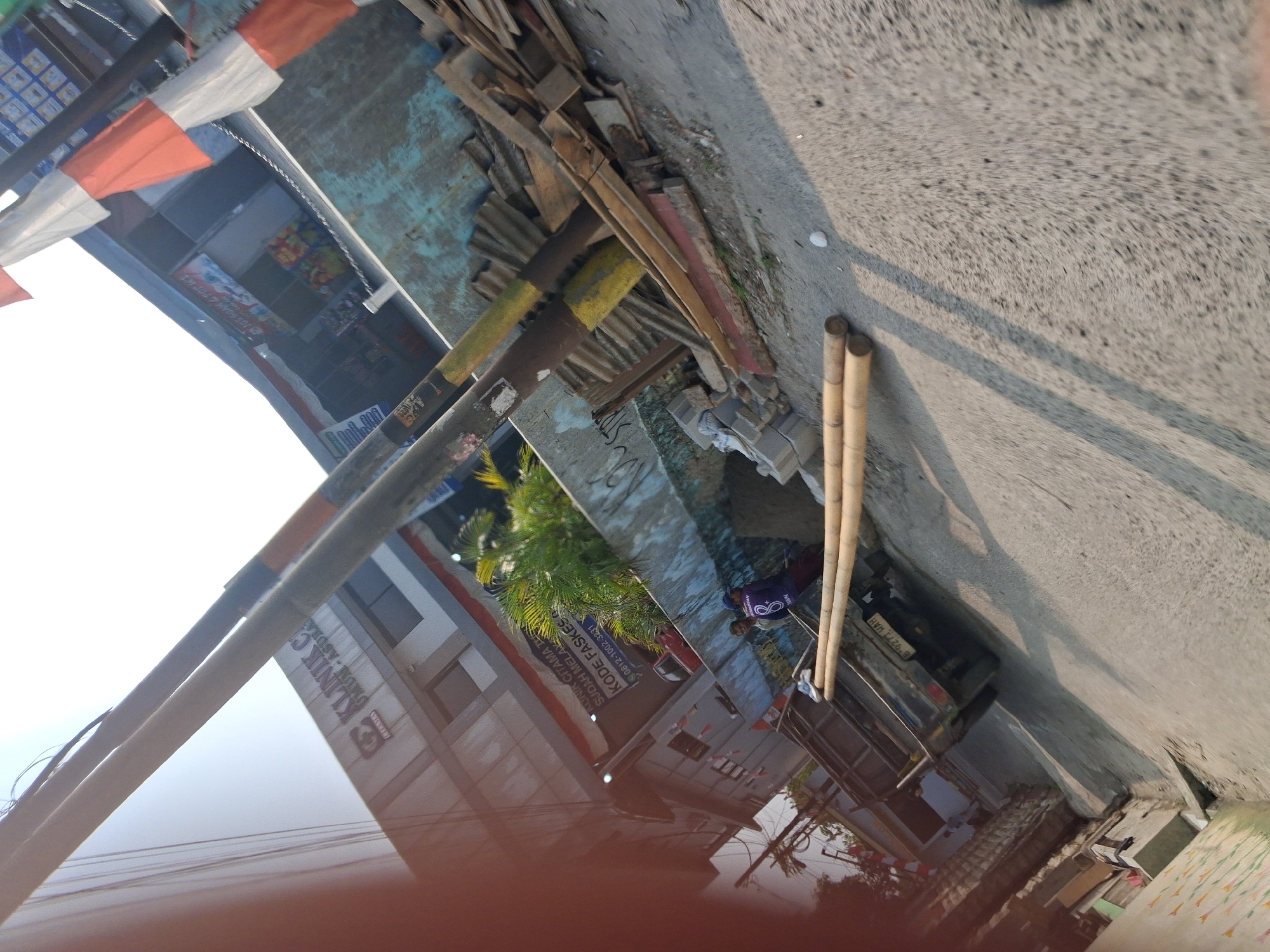 The image, taken in Bojong Gede, Indonesia, depicts a street scene with several elements. In the foreground, two prominent bamboo poles lie horizontally across the asphalt road. A dark-colored pickup truck, with its tailgate down and license plate visible as "B 9271 HAH," is parked on the right side of the road. In the background, a building with signage that reads "KLINIK CITAMA TE" is visible. To the left of the clinic, there is a smaller shop with colorful products displayed inside. Two individuals are present near the building, one wearing a blue shirt with the number 8 on the back and another person in a darker shirt. The environment appears to be an outdoor urban setting, with a clear sky and shadows cast by the bamboo poles and other structures. There are also piles of wood and corrugated metal sheets near the building, suggesting construction or debris.