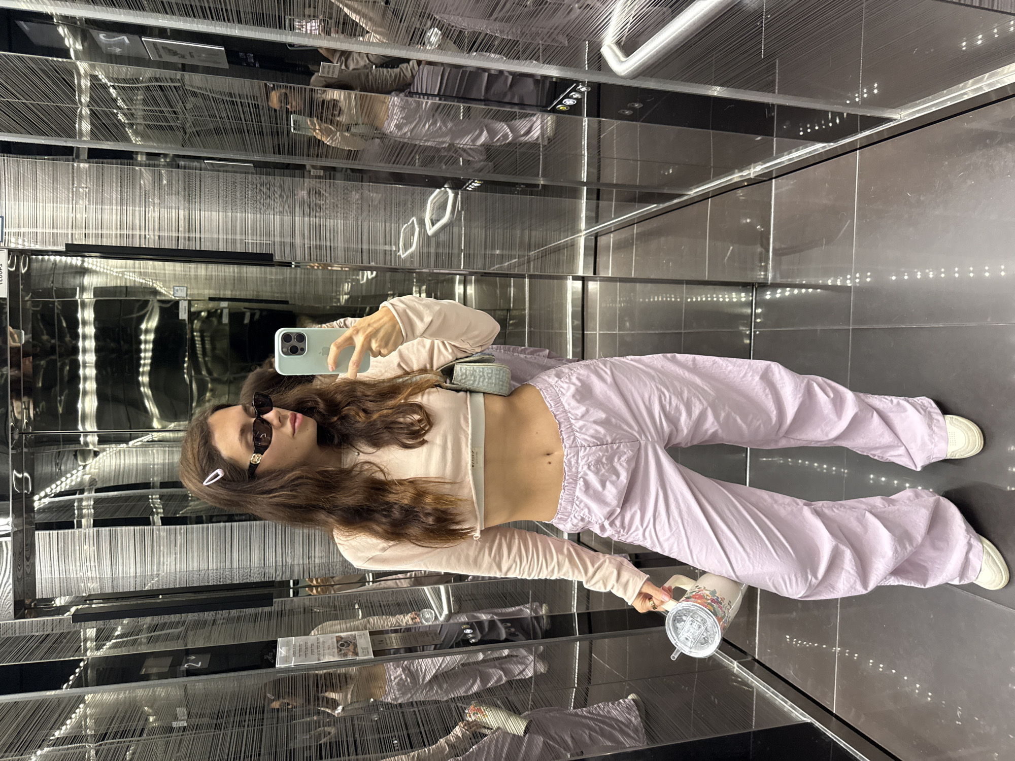 A young woman is taking a selfie in an elevator in Tel Aviv, Israel. She is wearing sunglasses, a light pink crop top and matching track pants, and white sneakers. She is holding a phone in one hand and a water bottle in the other. The elevator has mirrored walls and a unique design with vertical strands and metal accents.