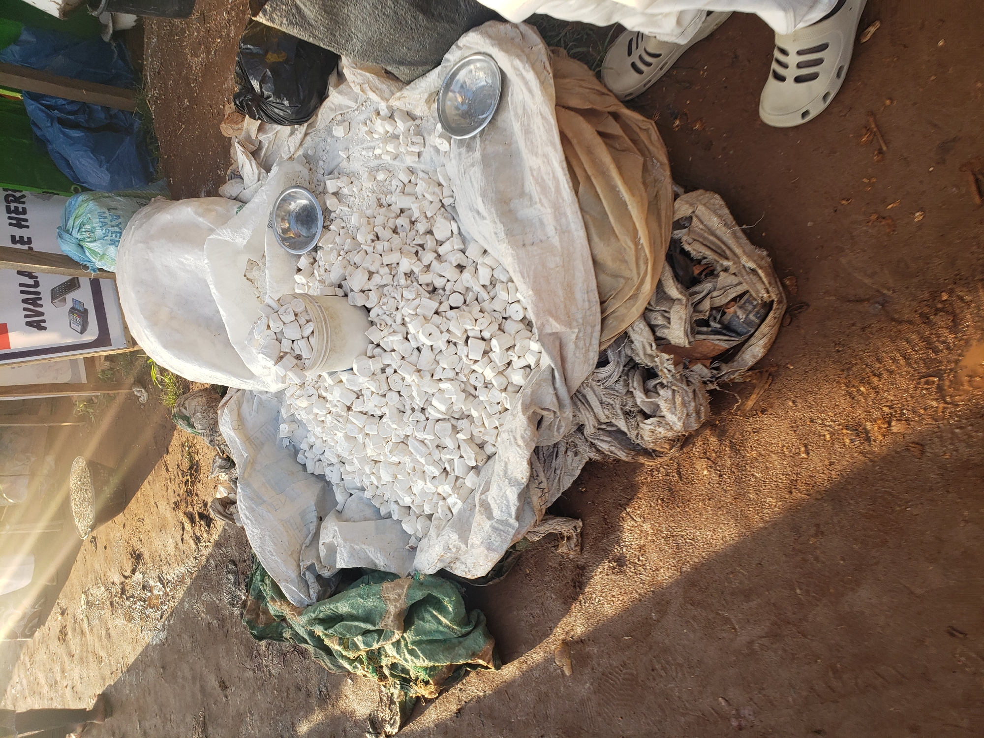 In Kakuri Gwari, Nigeria, a street scene unfolds with goods laid out on the ground. A pile of white, irregularly shaped objects, possibly a type of food or building material, is the central focus. These objects are contained within a large, white sack, with some spilling out onto a beige tarp. A small metal bowl rests on the tarp, and another is positioned near the pile of white objects. To the left, a green and brown sack lies on the dirt ground, partially obscuring a section of the pile. Behind the main display, a black plastic bag and a blue tarp are visible. Sunlight streams from the left, casting long shadows and illuminating parts of the scene, indicating it is likely daytime. The ground is a mix of dirt and scattered debris. The lower right of the frame shows the feet of a person wearing white trousers and light-colored clogs, suggesting their presence at this location.