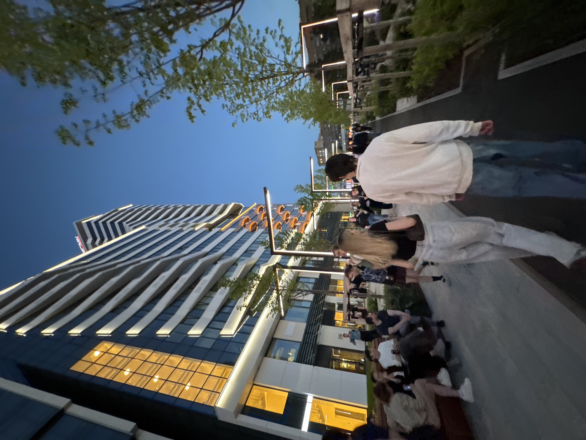 In Yekaterinburg, Russia, on a twilight evening, a group of people are gathered on a paved walkway adjacent to a modern, multi-story building with a glass facade. The building's exterior is illuminated with warm interior lights visible through its windows and accent lighting along its horizontal elements. Trees with green foliage line the walkway, interspersed with modern streetlights. In the foreground, two individuals are walking away from the camera on the walkway. To their left, a group of people are seated on a bench-like structure, engaged in conversation and holding drinks. The sky is a deep blue, indicative of the evening hour. The overall scene depicts a contemporary urban setting with a social atmosphere.