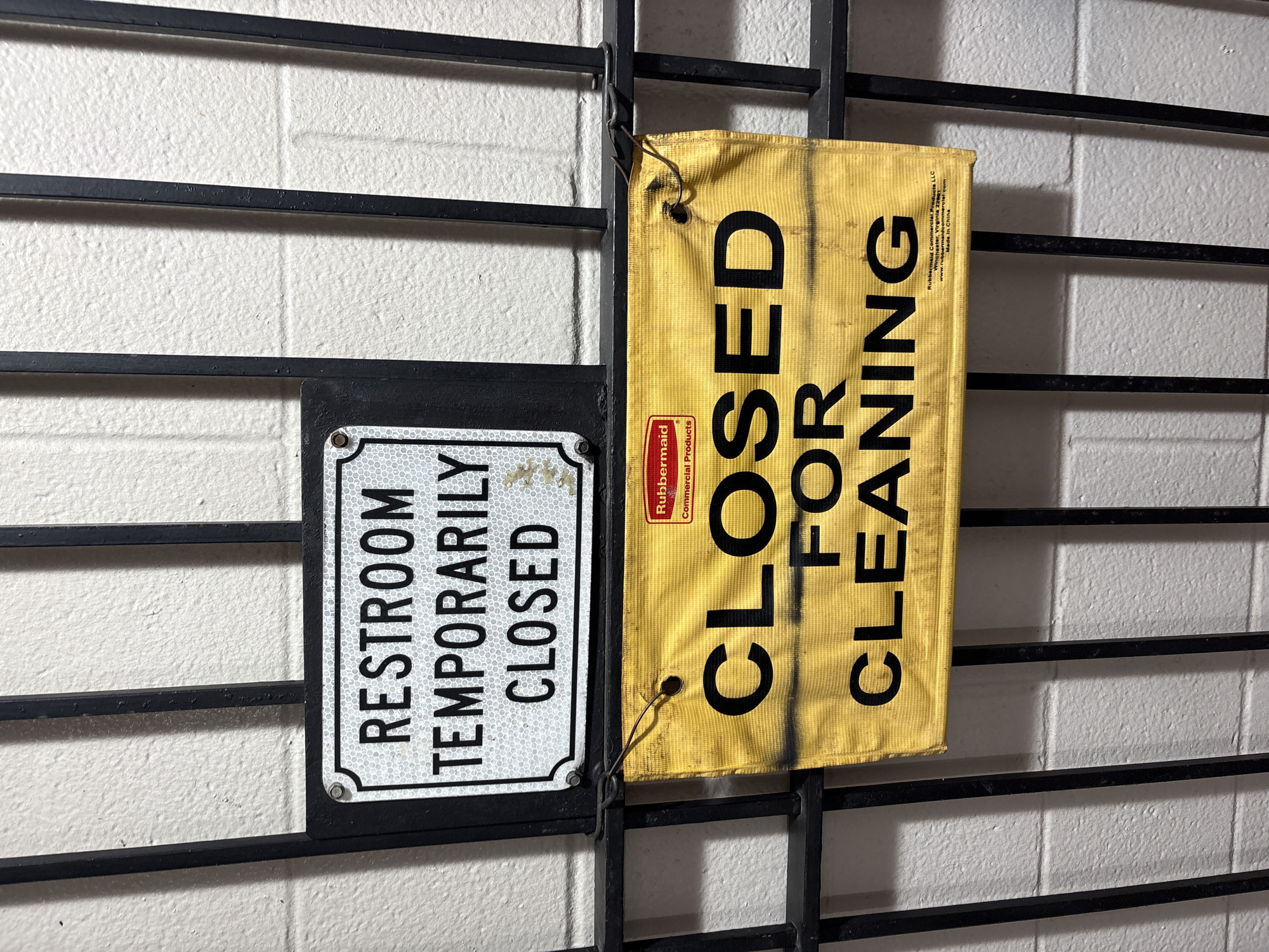 A close-up, eye-level shot shows two signs attached to a black metal gate in Sonora, California, United States. The gate is comprised of vertical bars. Behind the gate, a white cinder block wall is visible.
The upper sign is a white, rectangular plaque with a silver reflective border. It is attached to the gate with a screw at the top left corner. The text on the sign reads, in black sans-serif font, "RESTROOM TEMPORARILY CLOSED".
The lower sign is a yellow, rectangular banner with black text and a small red Rubbermaid logo in the top right corner. This sign is attached to the gate using thin wire. The banner is wrinkled and appears to be made of a fabric material. The black text on the banner is larger and bolder than the text on the upper sign. It reads, in three lines: "CLOSED FOR CLEANING". Beneath the word "CLEANING," smaller text indicates "Rubbermaid Commercial Products LLC, Winchester, Virginia 22601, www.rubbermaidcommercial.com, Made in China." The banner also has some smudges of black spray paint or dirt.