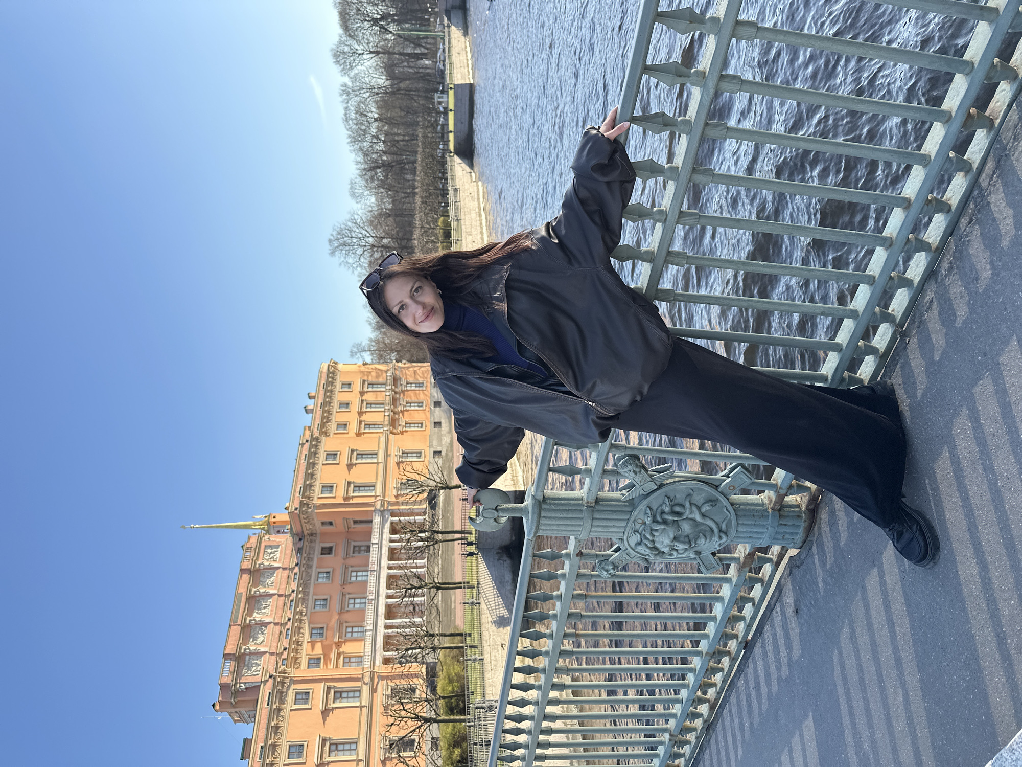 A portrait-style photograph, initially presented rotated 90 degrees counter-clockwise, captures a young woman posing on a bridge in Saint Petersburg, Russia, on a bright, sunny day. The woman, with long dark hair and sunglasses perched on her head, is wearing a dark blue turtleneck, a black leather jacket, and dark pants and shoes. She is smiling gently at the camera while leaning casually against a decorative, greenish-grey metal bridge railing. Her left arm extends along the railing, and her right hand holds onto an ornate bridge post featuring carved details resembling a Medusa-like head and winged figures.
To her left, behind the bridge, a wide body of water, likely one of Saint Petersburg's many canals or rivers, reflects the clear blue sky. On the opposite bank, several bare trees line the waterway, suggesting the season is either early spring or late autumn. Dominating the background is a large, majestic building with a light orange facade, adorned with numerous windows and intricate architectural elements, including a distinctive golden spire. This building strongly resembles the historic Mikhailovsky Castle (St. Michael's Castle) in Saint Petersburg. The sky above is a vibrant, cloudless blue, indicating clear weather and bright daylight, as evidenced by the strong shadows cast on the bridge. The woman's relaxed posture and pleasant smile convey a serene and happy mood. There is no visible text in the image.